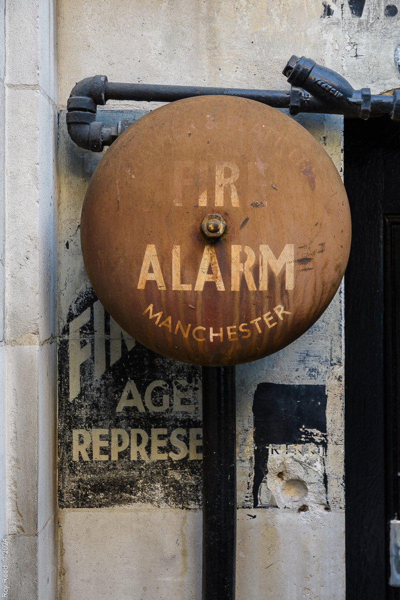 A large rust-red fire alarm bell with a faded painted sign behind it.