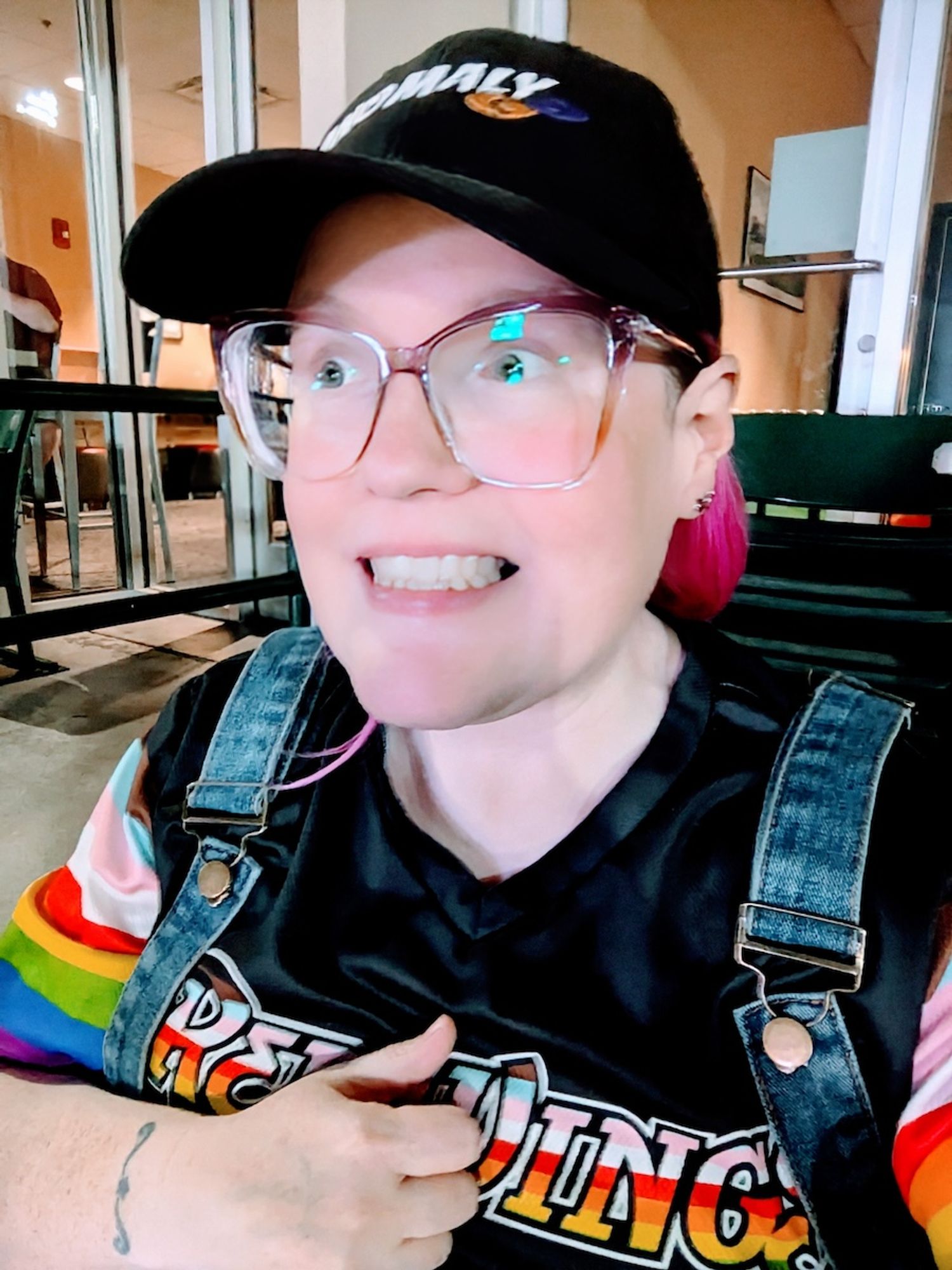 Self-portrait at the ballpark. Wearing a black Anomaly baseball hat with some pink hair poking out, a Pride style Red Wings jersey under overalls, and big frame glasses. My expression is a worried, toothy smile as I watch a fly ball getting worrisomely close.