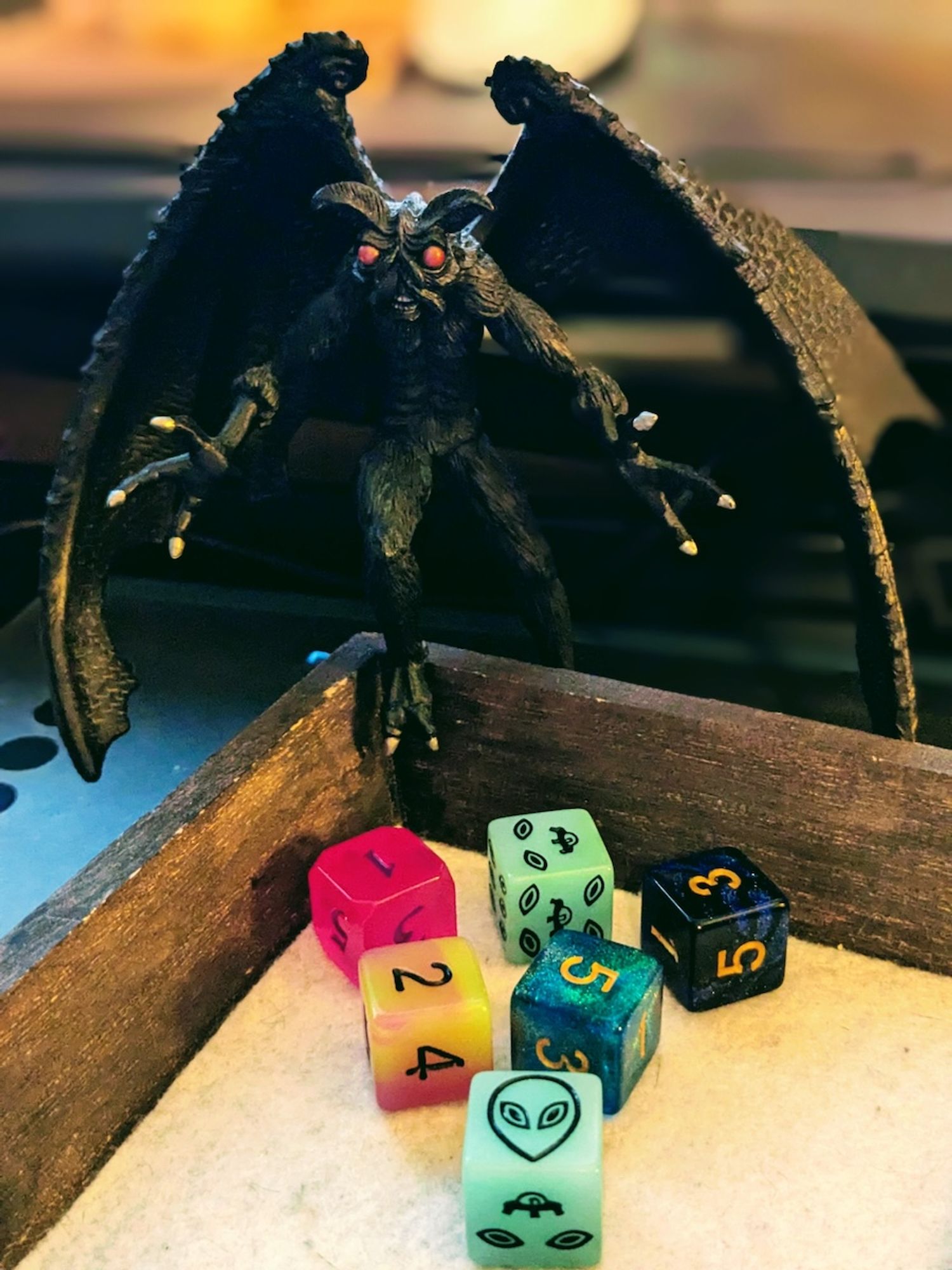 A pile of six 6-sided dice in a wooden tray, with a Mothman figurine looming above them.