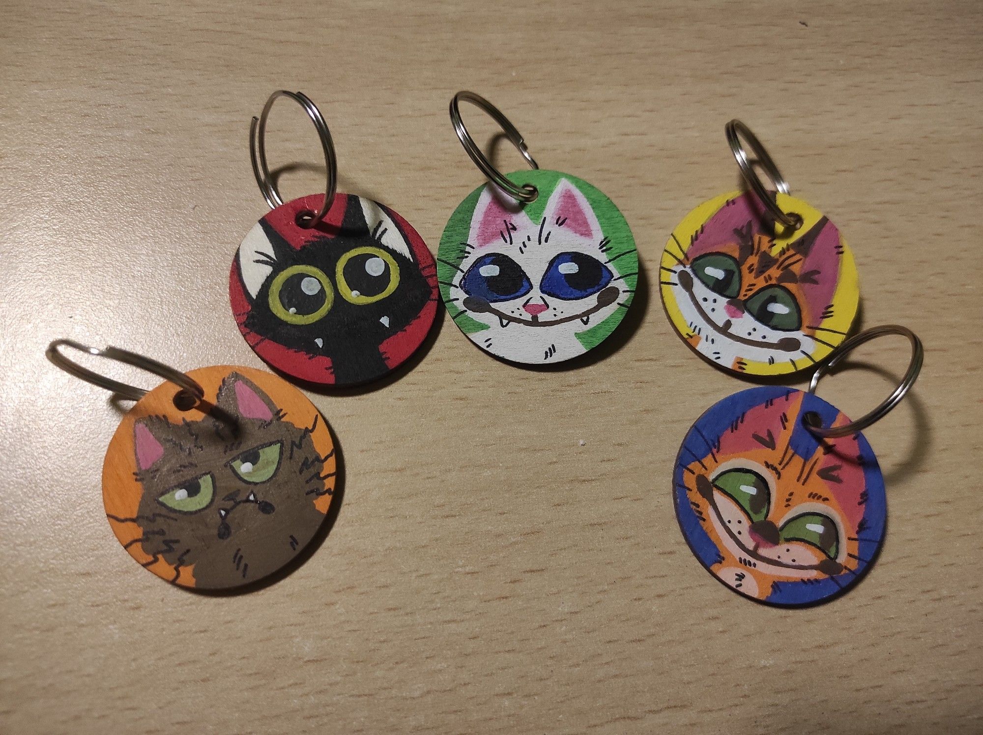 Five round, wooden charms with (left to right)
- a brown, fluffy frowning cat with green eyes 
- a black void Cat with big yellow eyes and pointy teeth 
- a smiling white cat with blue eyes
- a smiling orange and white cat with stripes and green eyes 
- a smiling orange with cream cat with green eyes