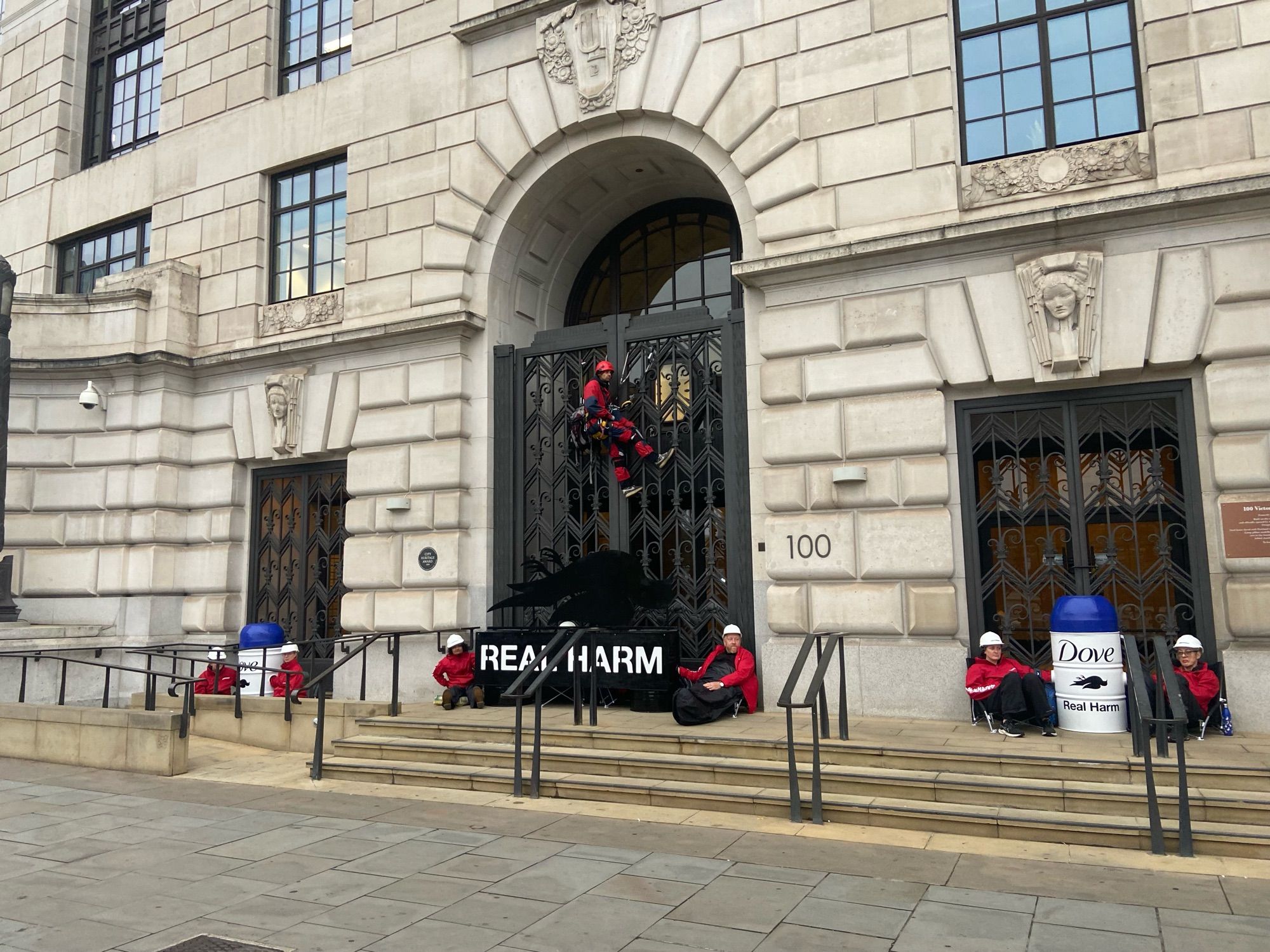 A photo of Greenpeace protesters dressed in red, locked to the entrance to the Unilever office building in London. The protesters are chained to the gates and have a sign reading Real Harm.