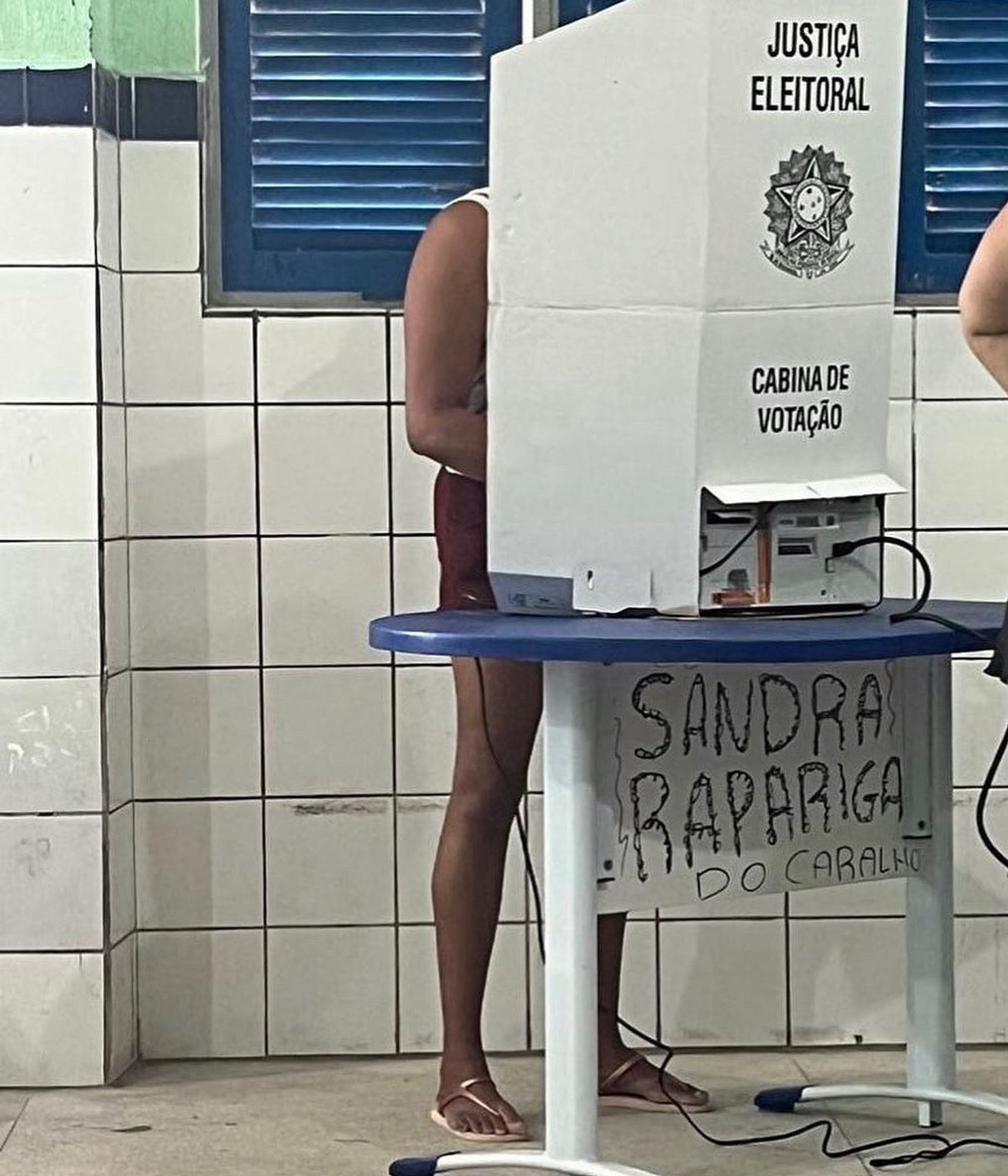 Imagem de urna eletrônica em uma escola. Na carteira onde foi colocada, alguém pichou: "SANDRA RAPARIGA DO CARALHO"