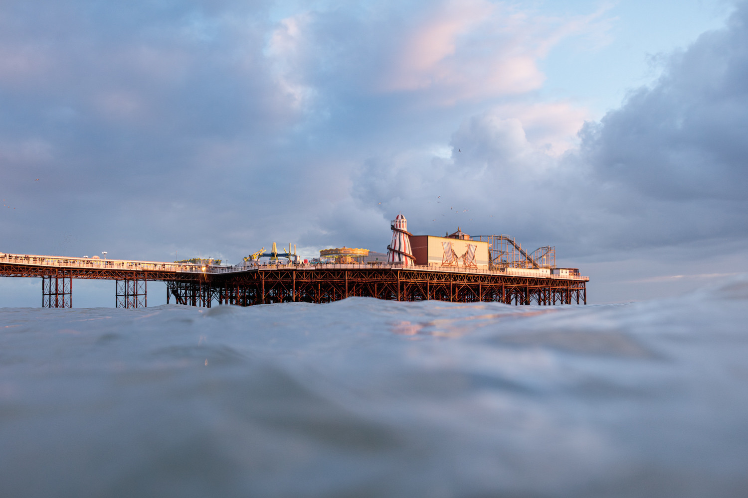 Brighton Palace Pier as seen from the sea. The pier is lit in warm winter light under a moody, cloudy sky. 