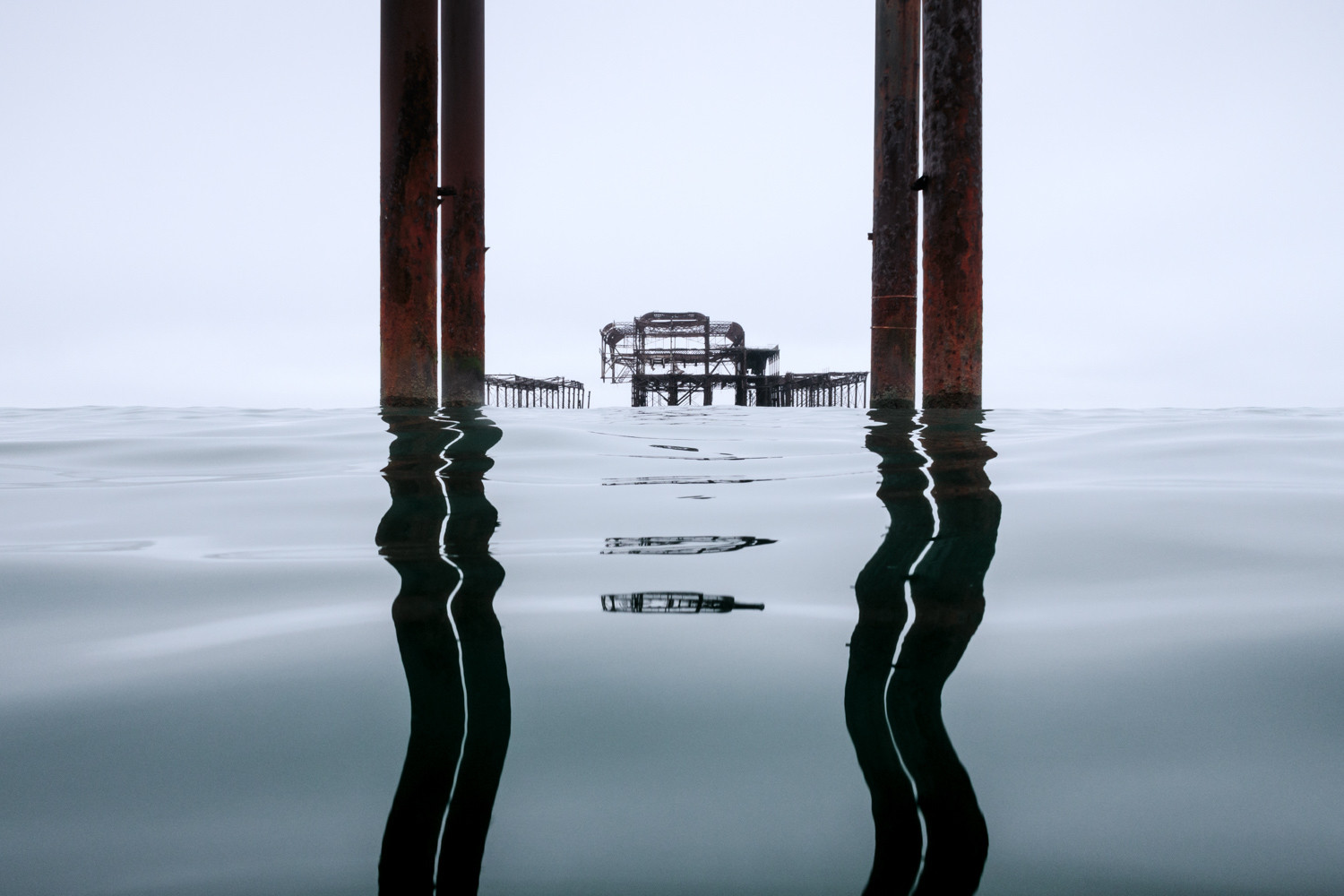 Brighton's West Pier as seen from the sea. The pier is framed between the tall iron columns that stand in front of the pier, their reflections wiggle neatly on the waters surface. 