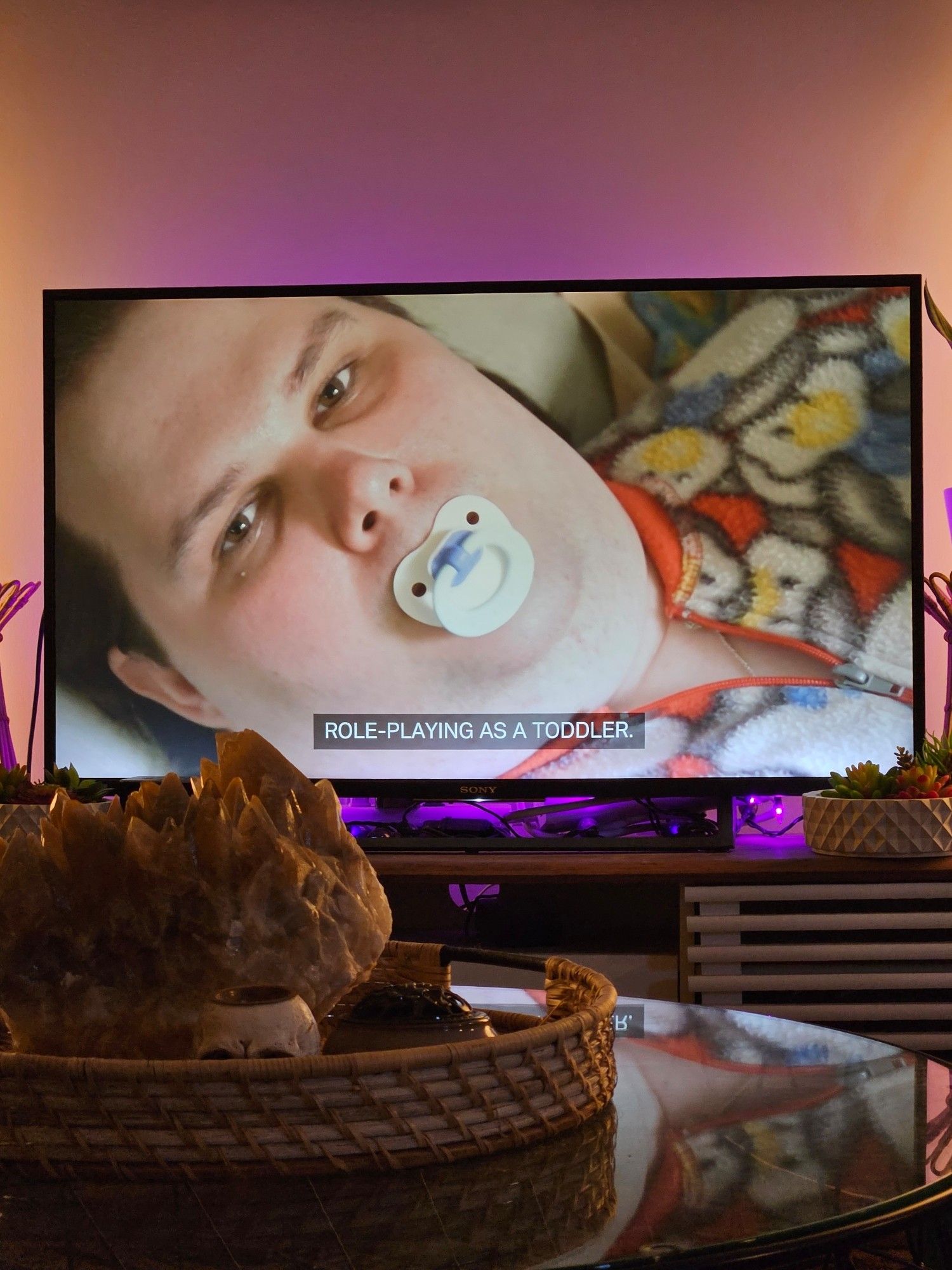 My tv showing an adult male with a baby pacifier. Closed captions text says "role-playing as a toddler"