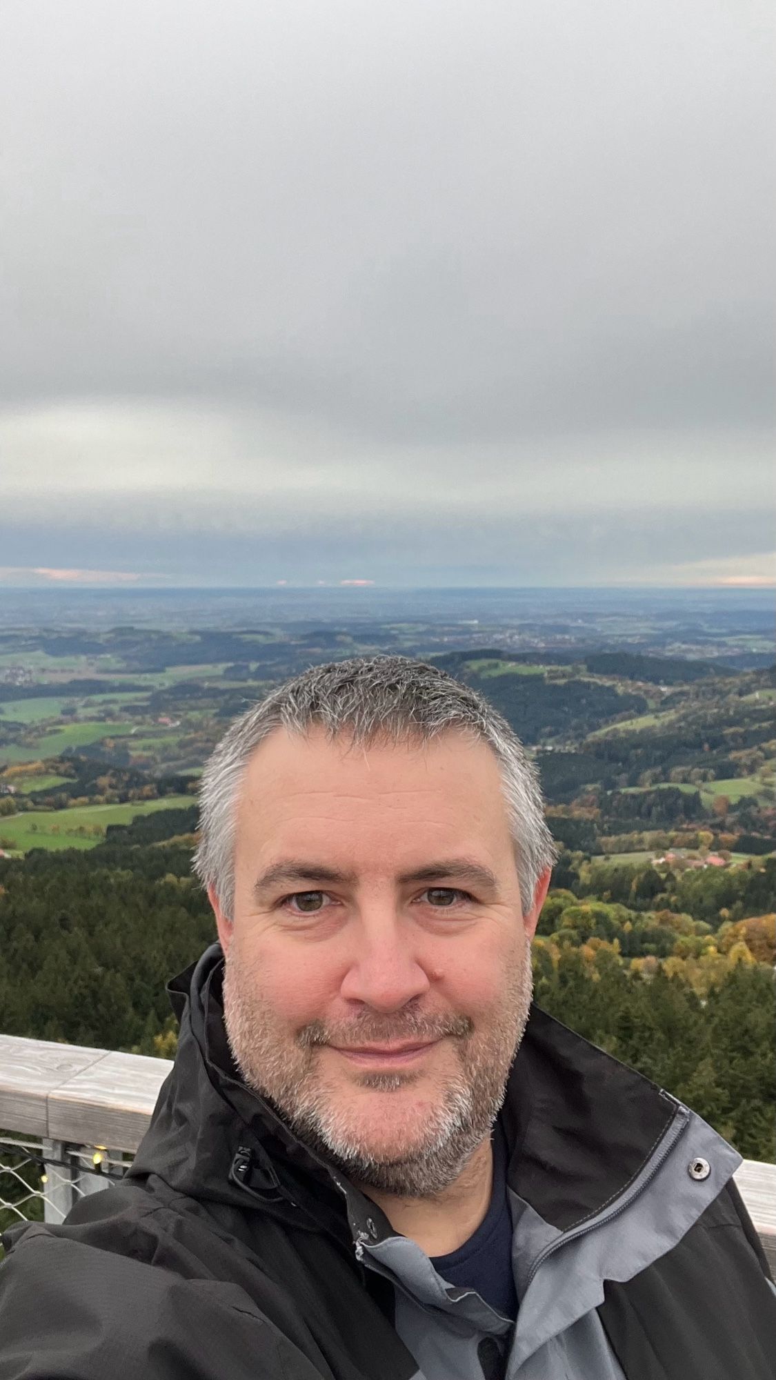 Ich stehe auf dem Turm des Waldwipfelweges St. Engelmar und schieße ein Selfie. Hinter mir ist ein tolles, aber wolkenverhangenes Panorama Bayerns zu sehen.