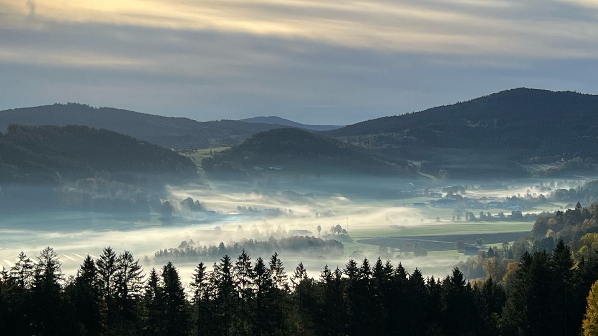 Die Sonne geht auf, im Tal hinter den Wiesen und Wäldern sind weiße dichte Nebelfelder zu entdecken