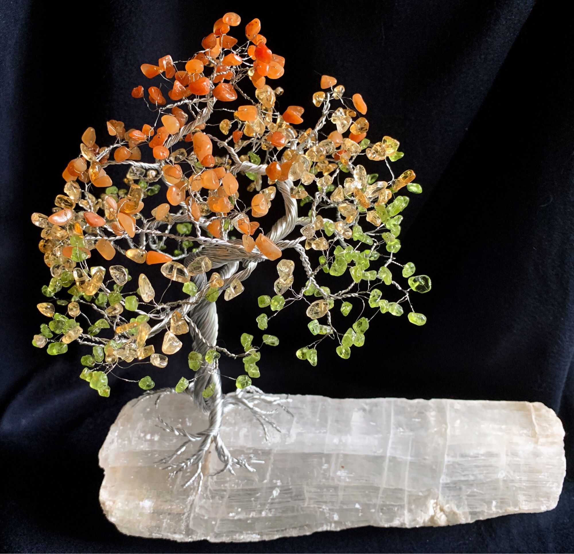 A silver wire tree with green, yellow, and red stones for leaves, mounted on a piece of selenite and displayed on a black background