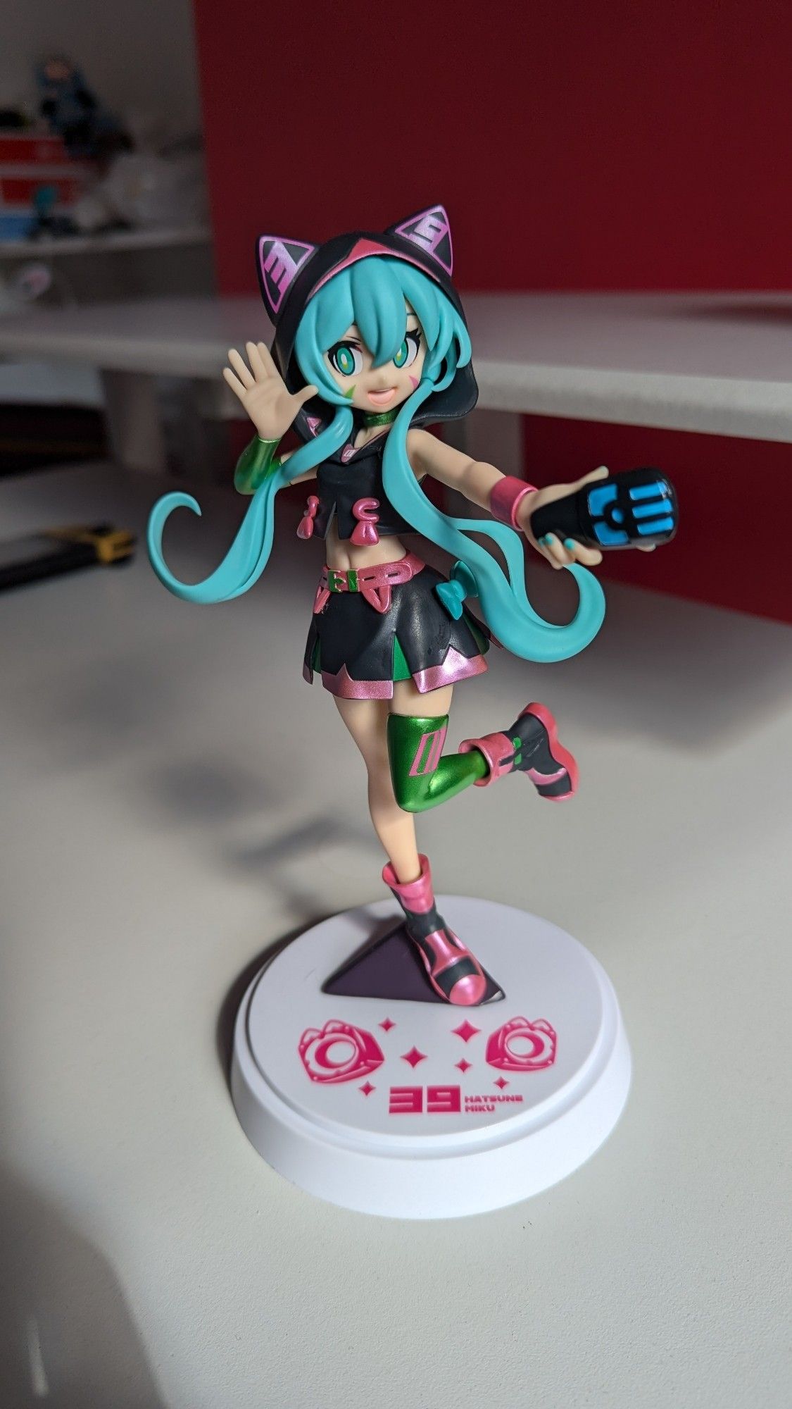 A Hatsune Miku figure. She is in an active pose, with one leg up, an outstretched arm holding a microphone, and her other hand to her ear. She is wearing a cropped hoodie with cat ears, and a small skirt, both of which are black with red highlights. Her blue hair is flowing out and over her arms.