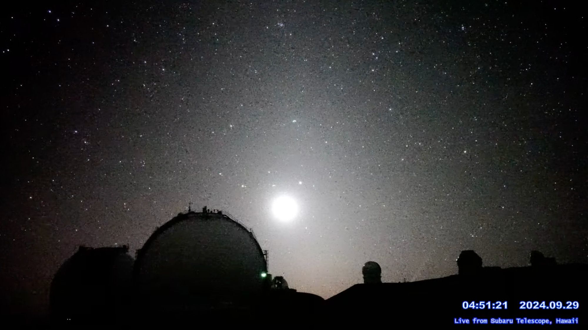 A webcam image showing outlines of dark observatory domes, many stars, the bright overexposed Moon and the line of the comet's tail between two domes at right.