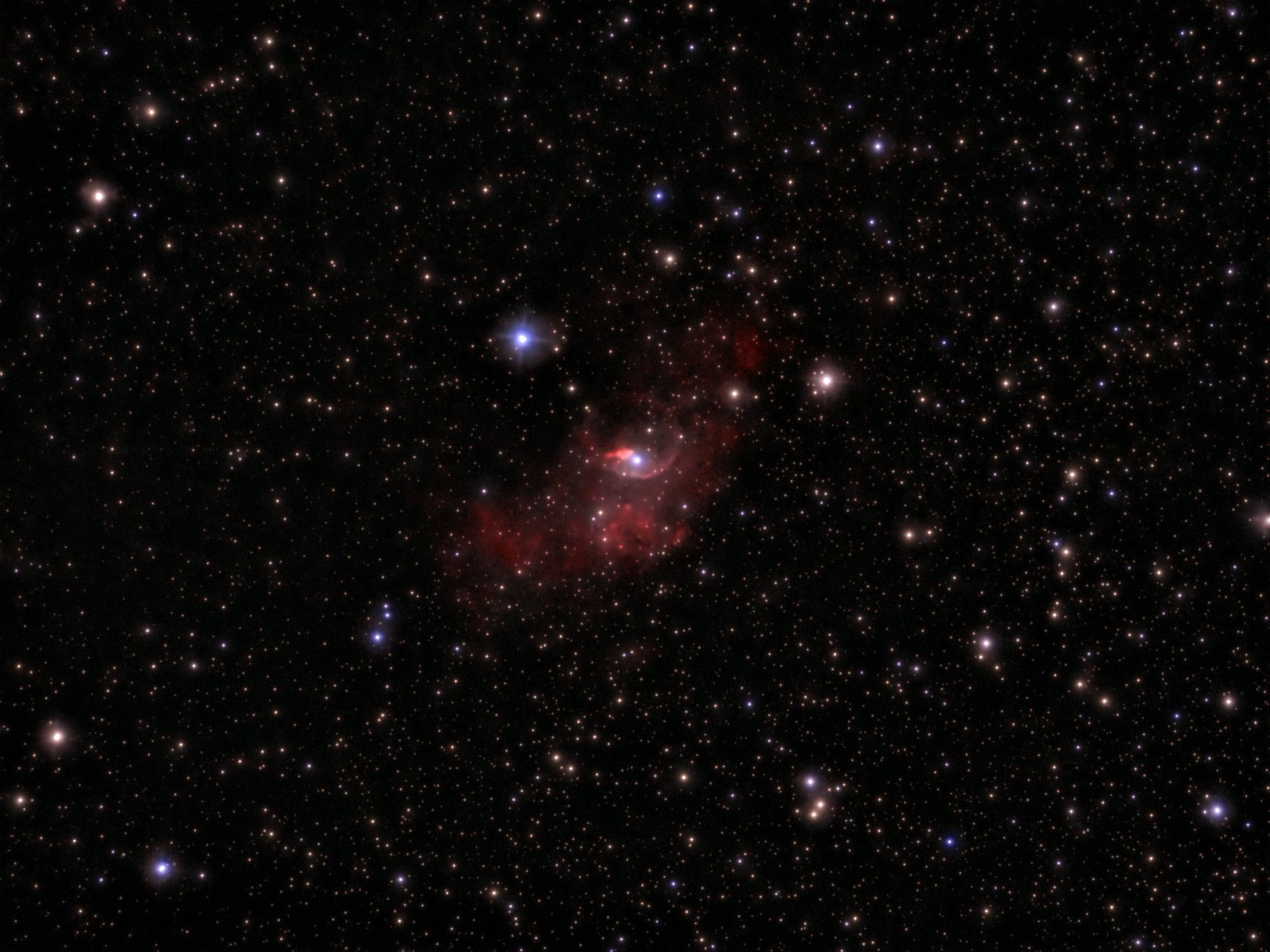 A telescopic image of a field of stars against the blackness of space. The stars show different colors. At center is a red cloud with a bubble of gas that has been blown out from the bluish star at the center.