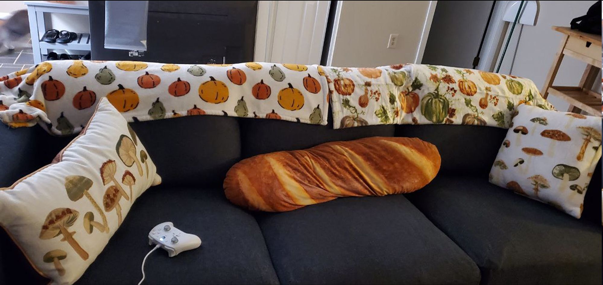 Picture of a dark grey couch with 2 mushroom designed throw pillows, two pumpkin themed throw blankets, and a large baguette pillow in the center. Also an xbox controller, just cuz