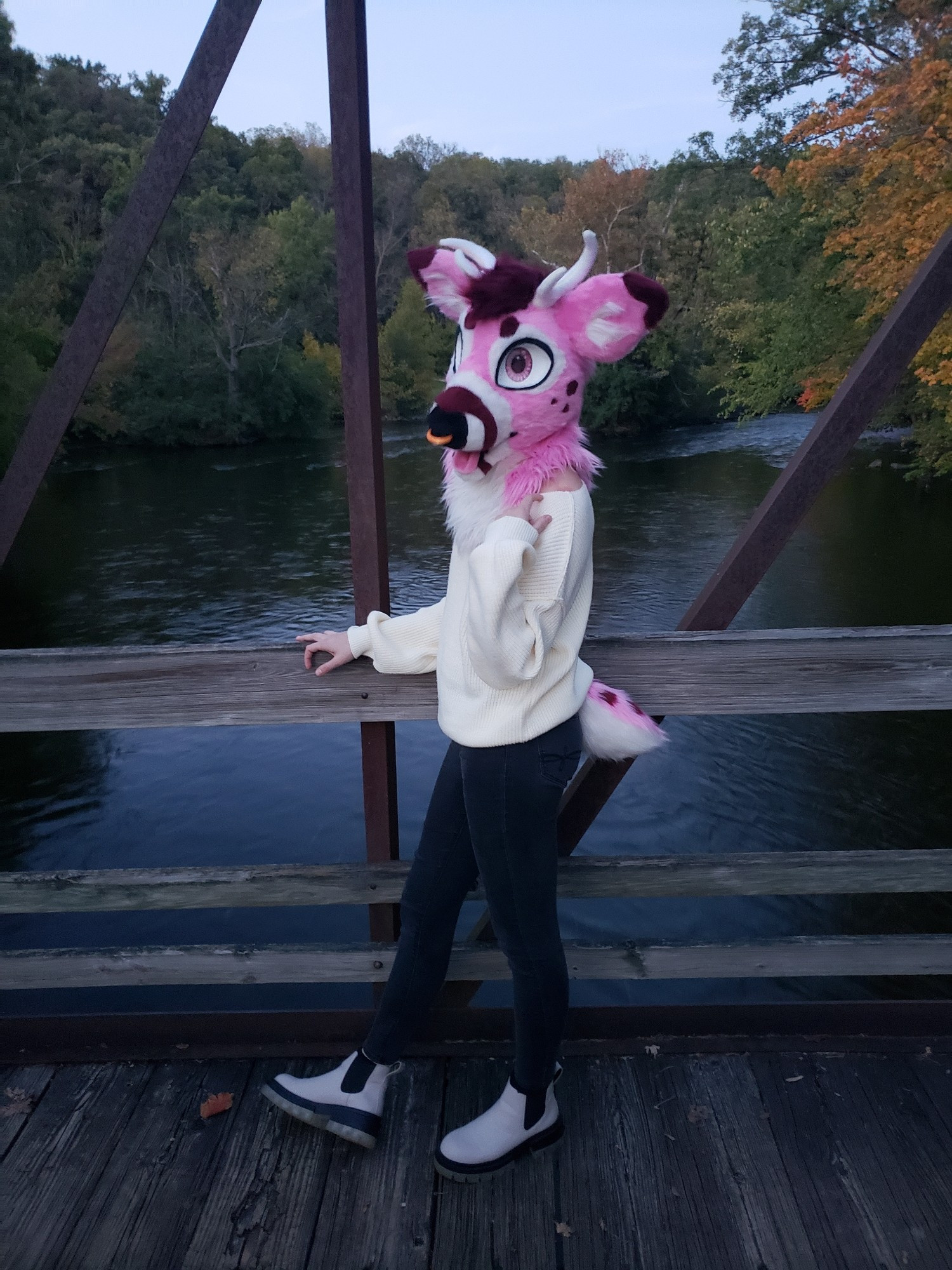 Pink deer Tavi on a bridge. Her arm is raised up to her shoulder. The background is a river and woods with some of the leaves starting to turn color