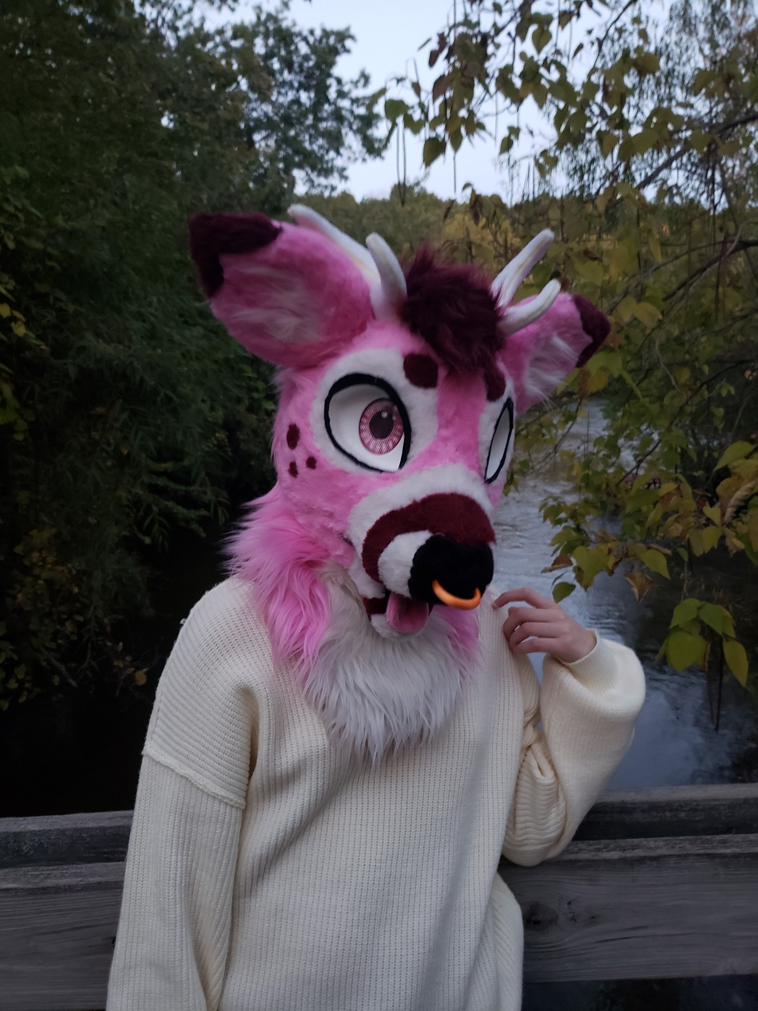 Pink deer Tavi standing on a bridge in a sweater. She has one hand raised to her shoulder