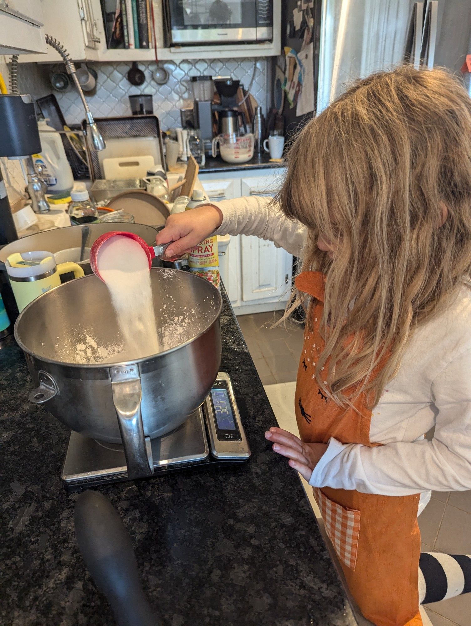 Young blond haired girl wearing an orange apron with a cat face on it pouring sugar into a metal bowl on a scale that reads 321g. The sink in the background is full but the counter is clean.