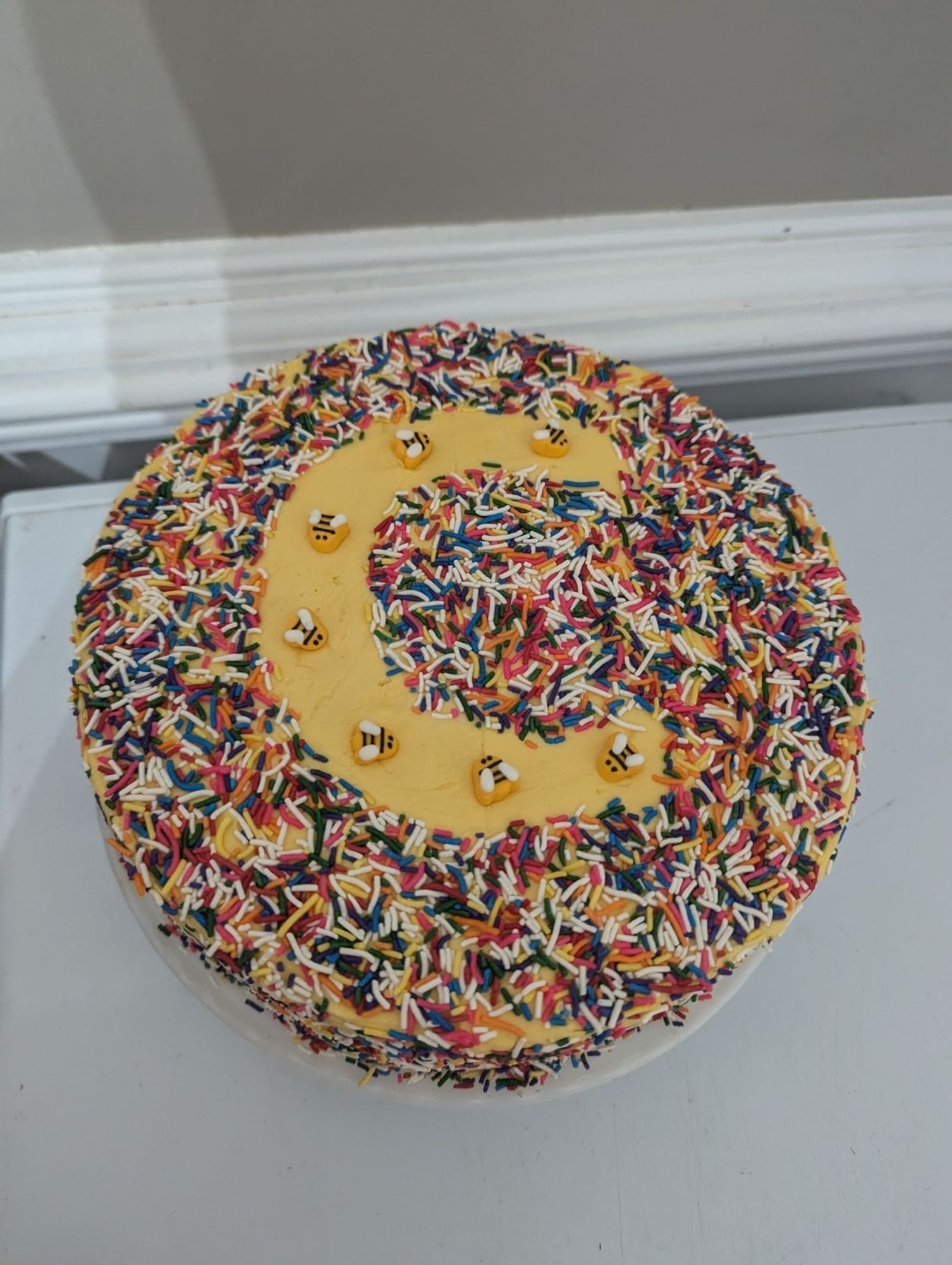 A cake with yellow frosting, covered with rainbow sprinkles, and a giant "C" in the negative space with 7 candy bees, on a chest freezer next to a gray wall