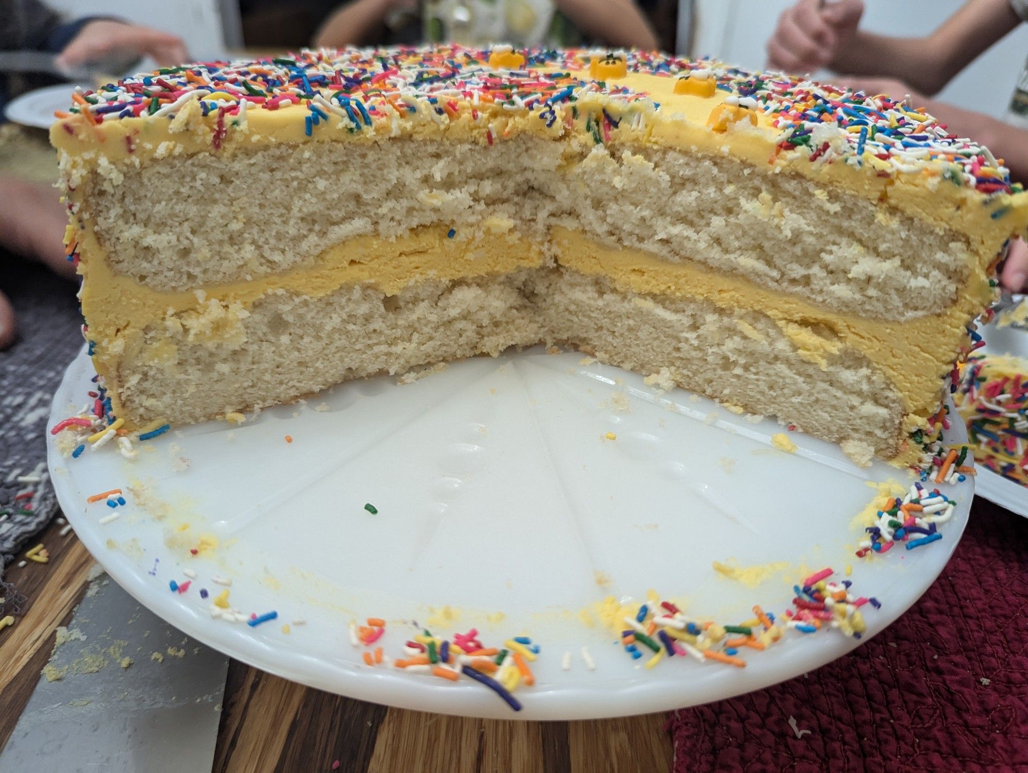 Cross section of a 2-layer white cake with yellow frosting and rainbow sprinkles, on a white cake stand on a bamboo table. Kids' arms are blurred in the background as they devour the cake.