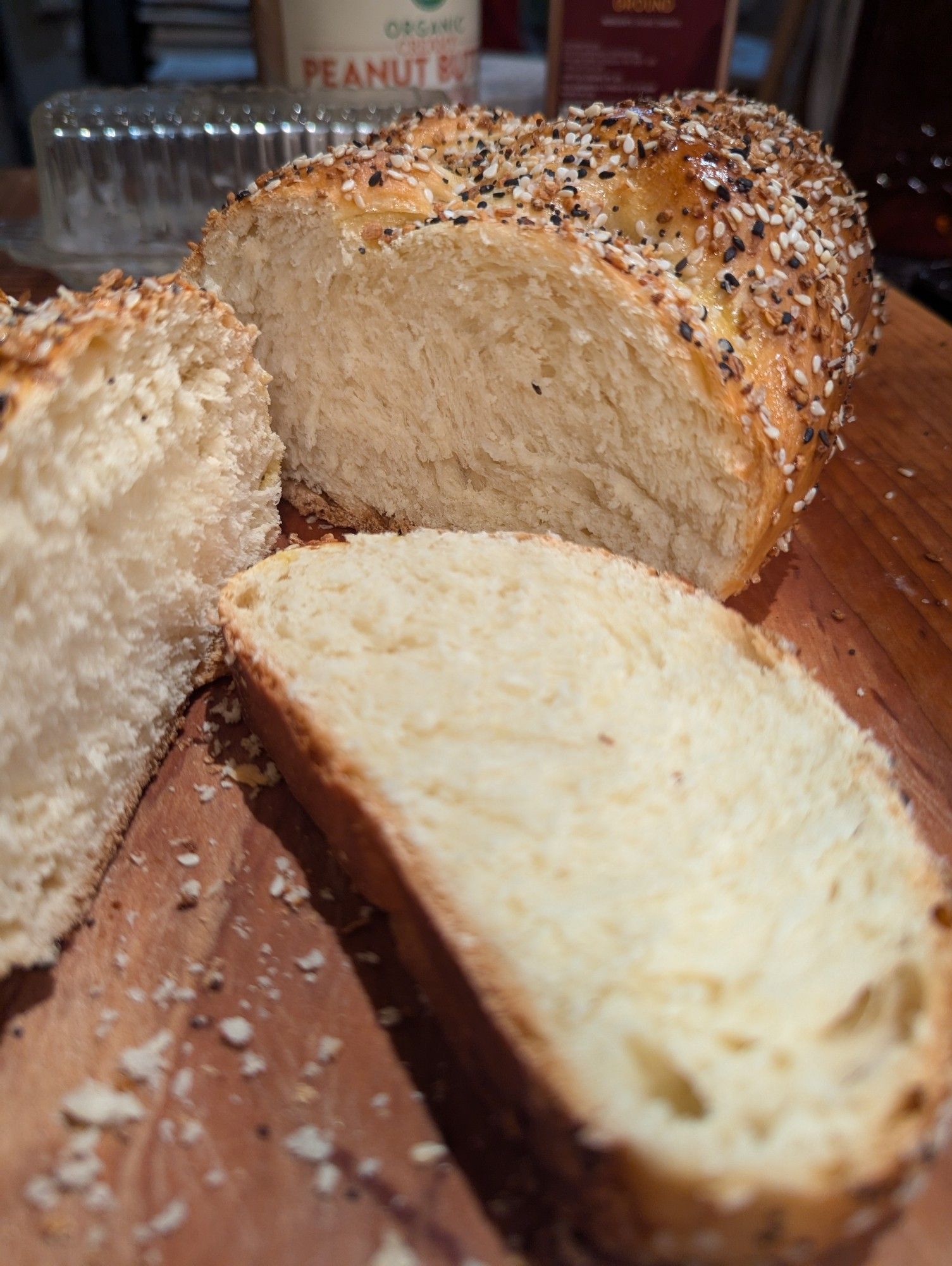 Cross section of a 3-strand challah topped with everything bagel seasoning, on a wooden cutting board, blurry butter dish and peanut butter jar in the background