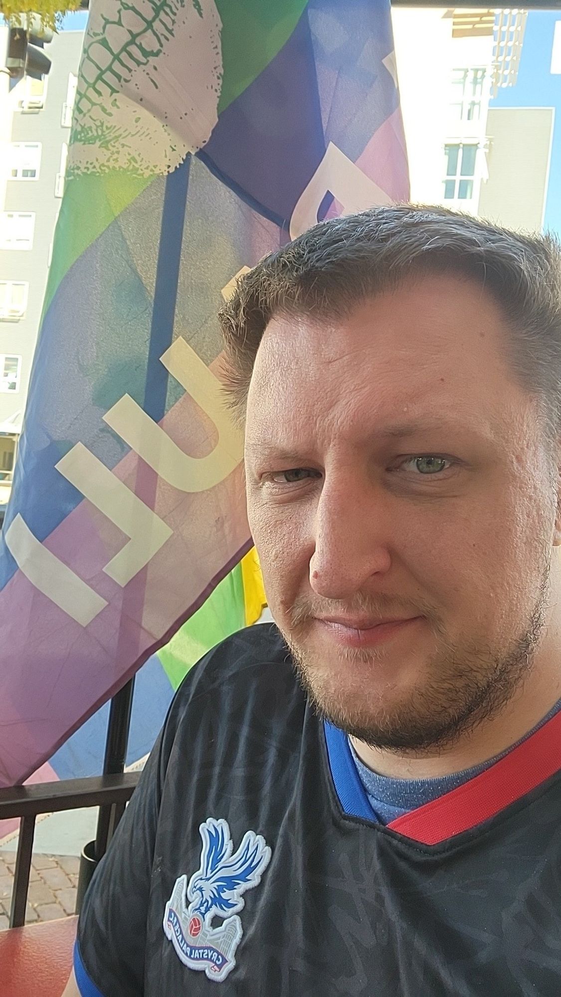 A white man wearing a crystal palace shirt and sitting next to a pride flag with the St. Pauli FC logo on it