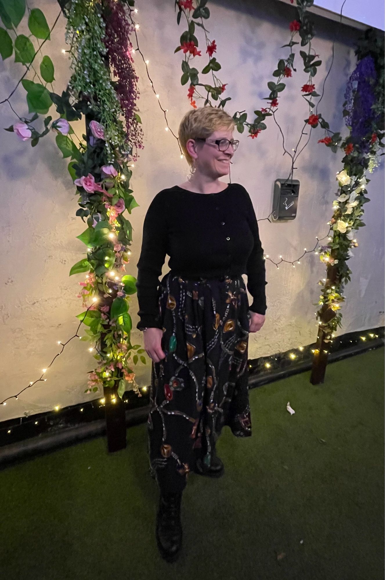 I’m standing in front of a wall with fake plants and fairy lights. Wearing a black cardigan and a long black skirt with large jewels and strings of pearls as the pattern.