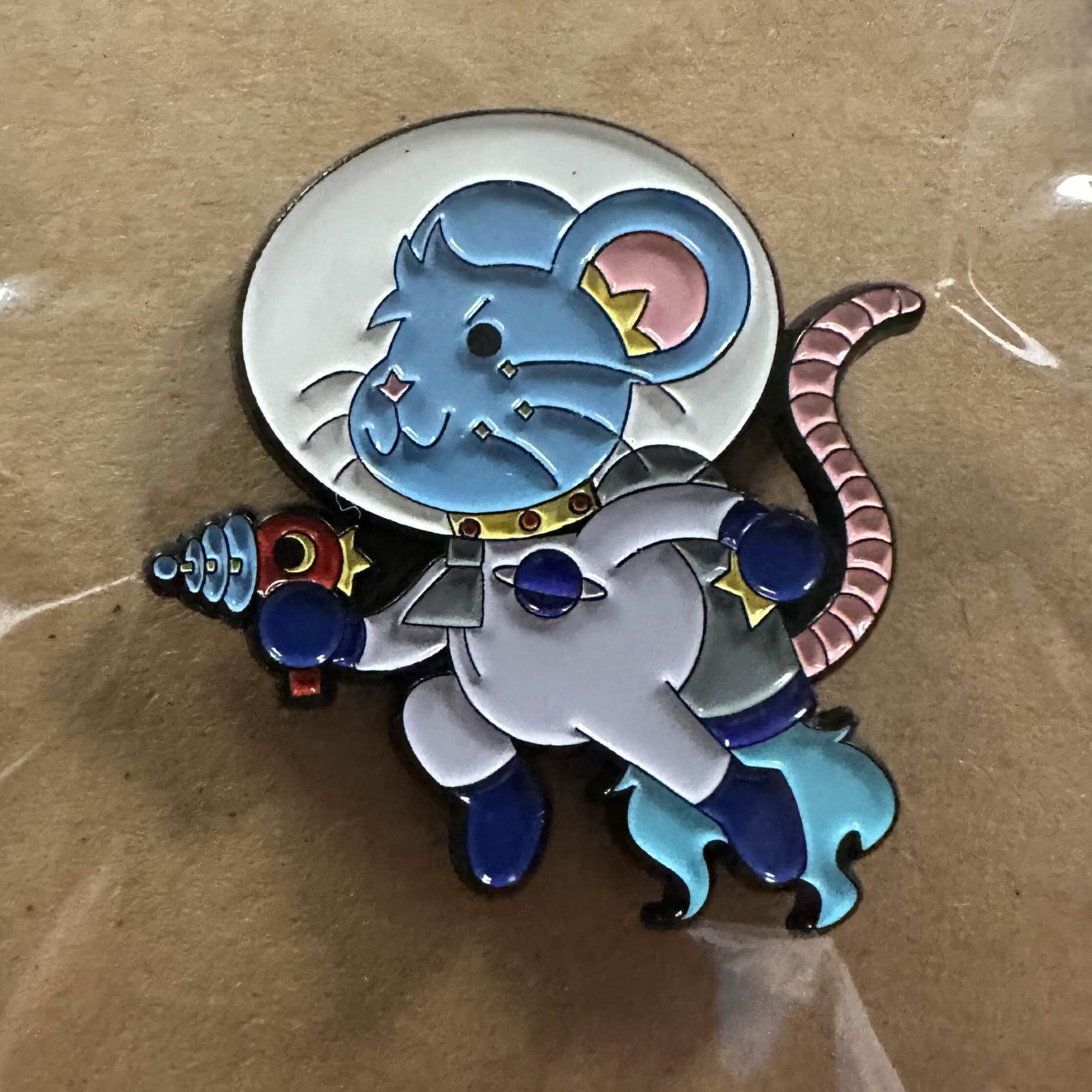 An enamel pin featuring a blue rat wearing a space suit, holding a ray gun and flying with his jetpack