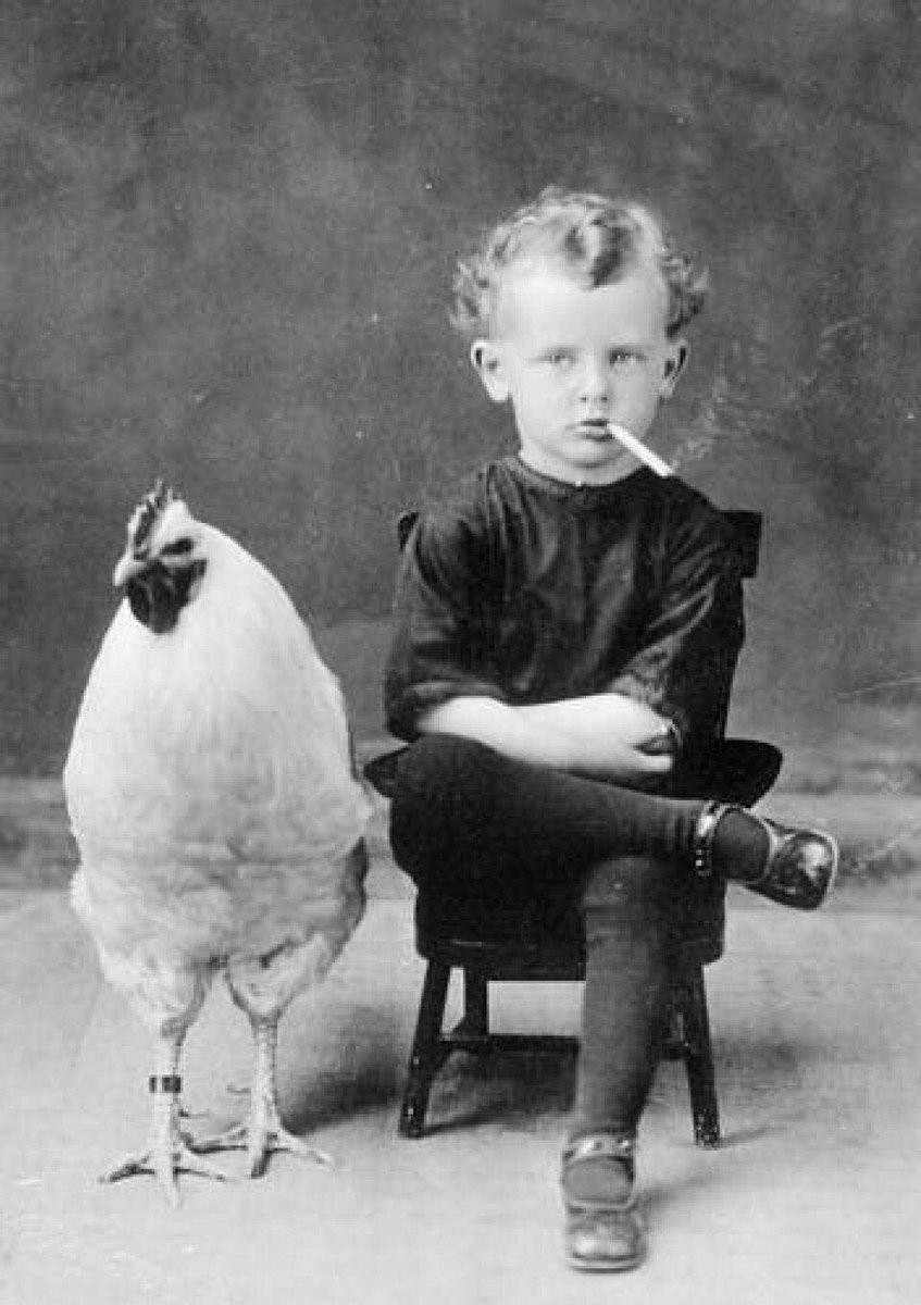 a old photo of a small boy sitting crosslegged in a chair while smoking a cigarette. A rooster stands nearby.
