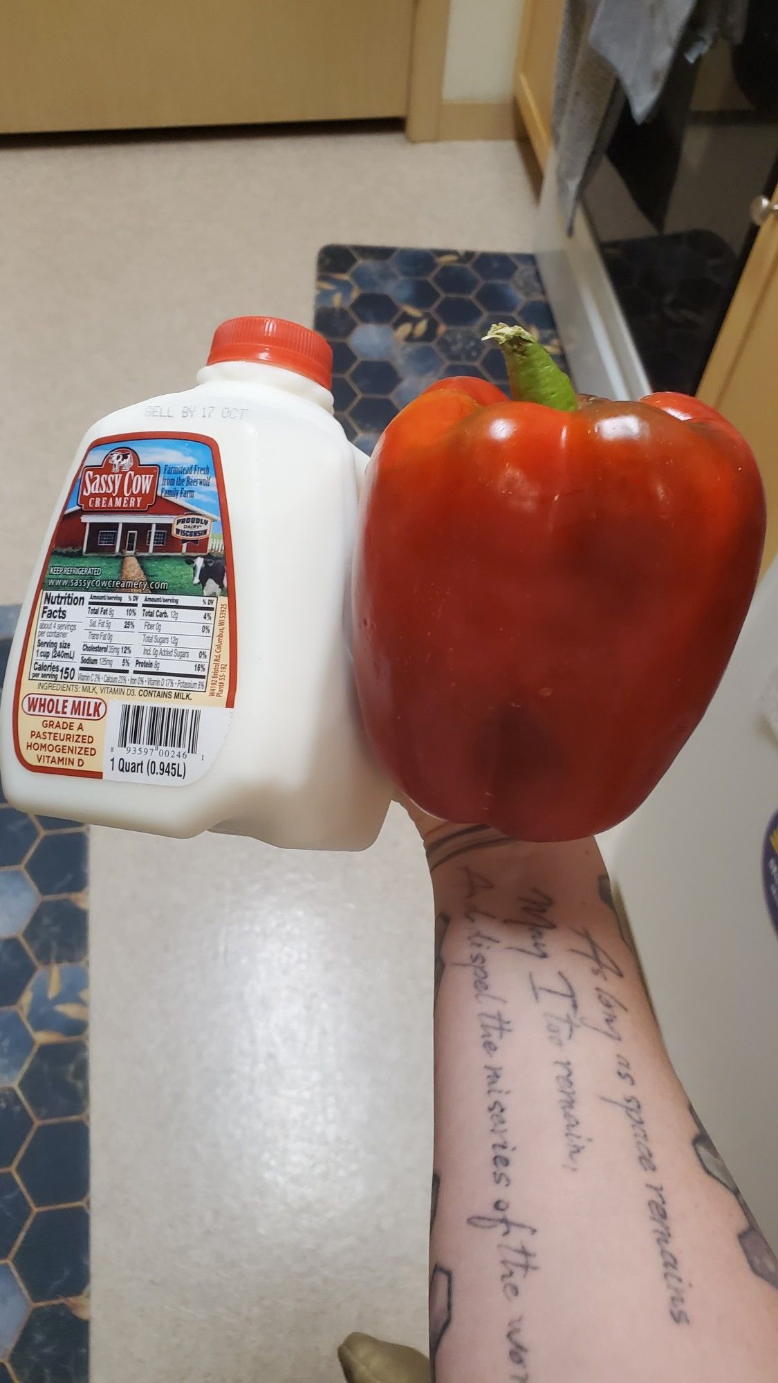 Sassy cow creamery milk, 1 quart next to a giant red bell pepper. About the same size. Being held by one hand of a white arm with tattoo.