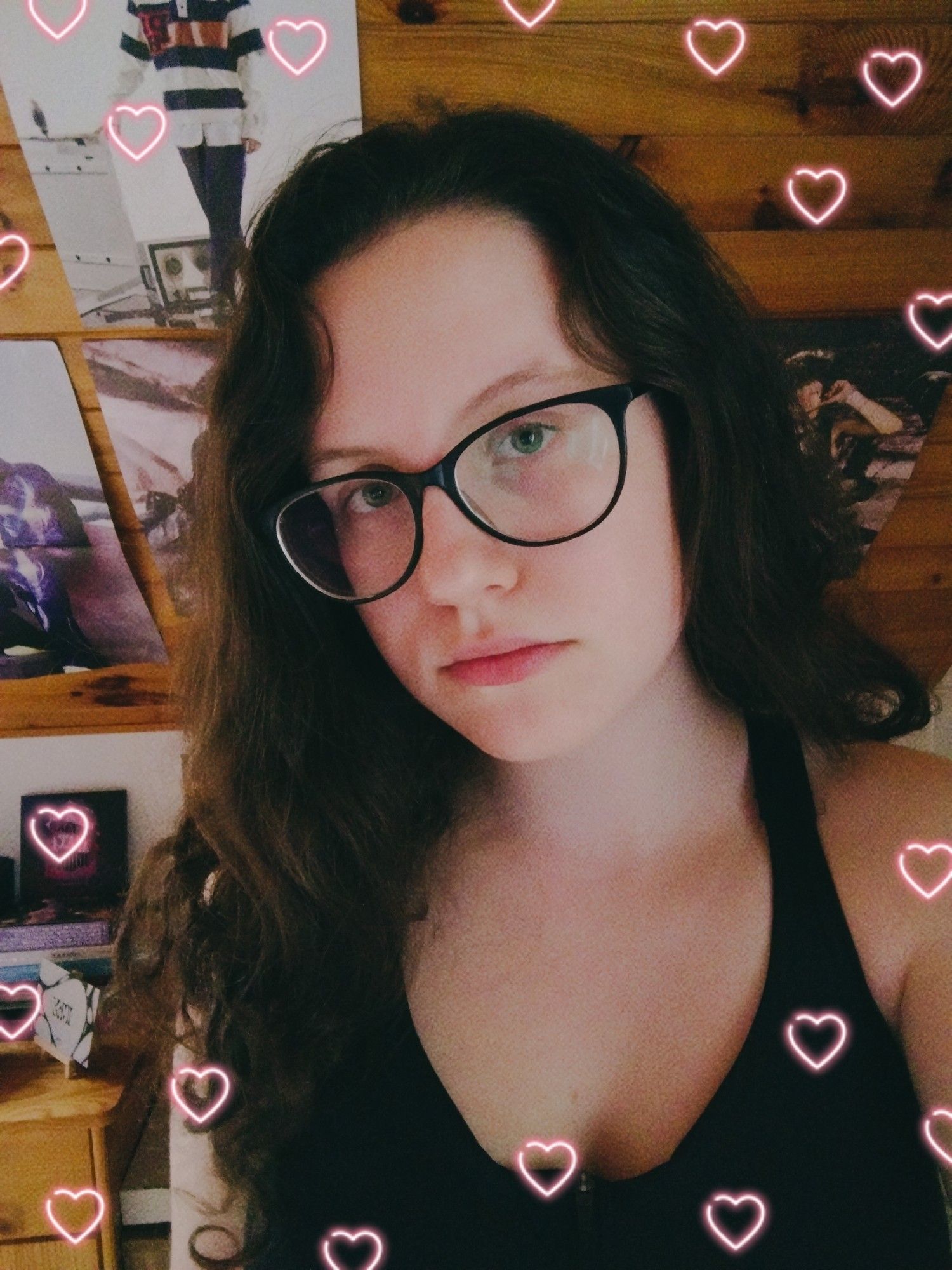 Just a selfie of me in my room. I have dark brown wavy hair, glasses and green eyes. I'm wearing black sports bra. In the background you can see some of my kpop posters and albums. I edited the photo and added neon hearts frame.