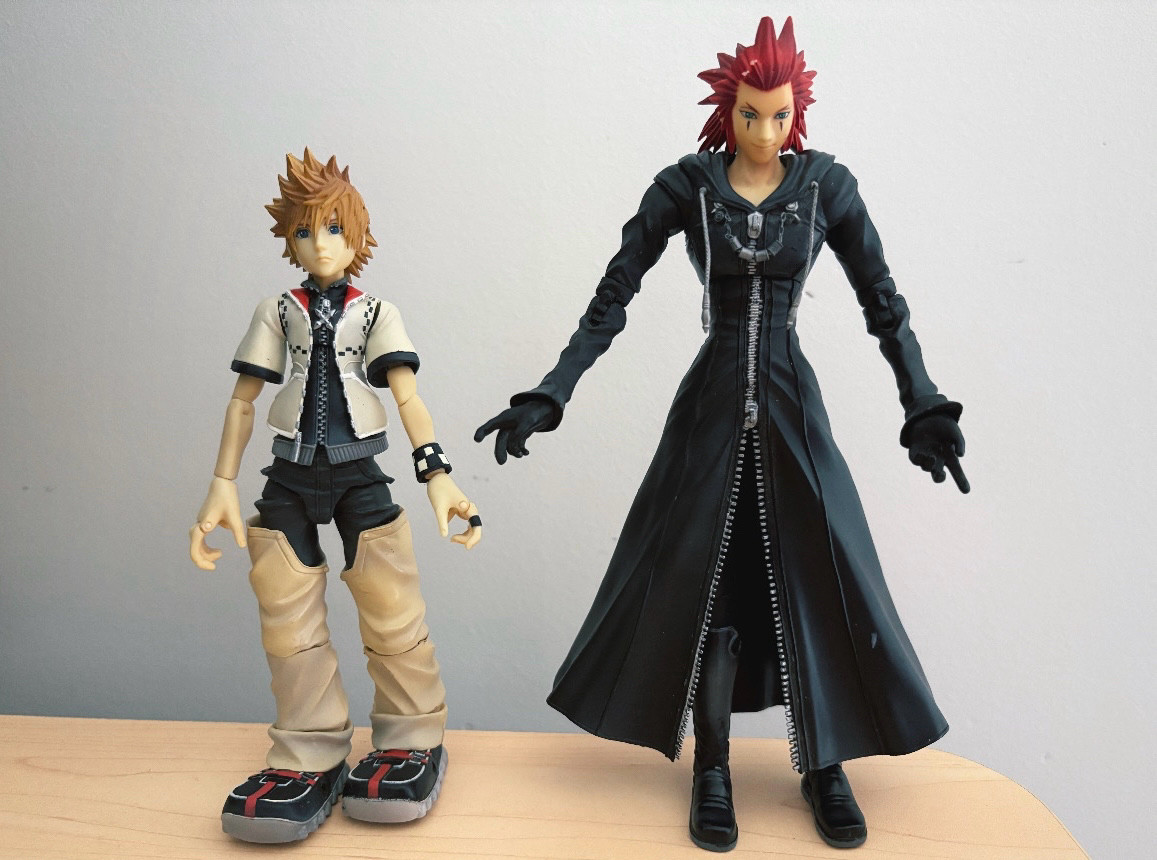 The Axel (right) and Roxas (left) action figures from the Kingdom Hearts II Play Arts series are set upright on a wooden bench. 