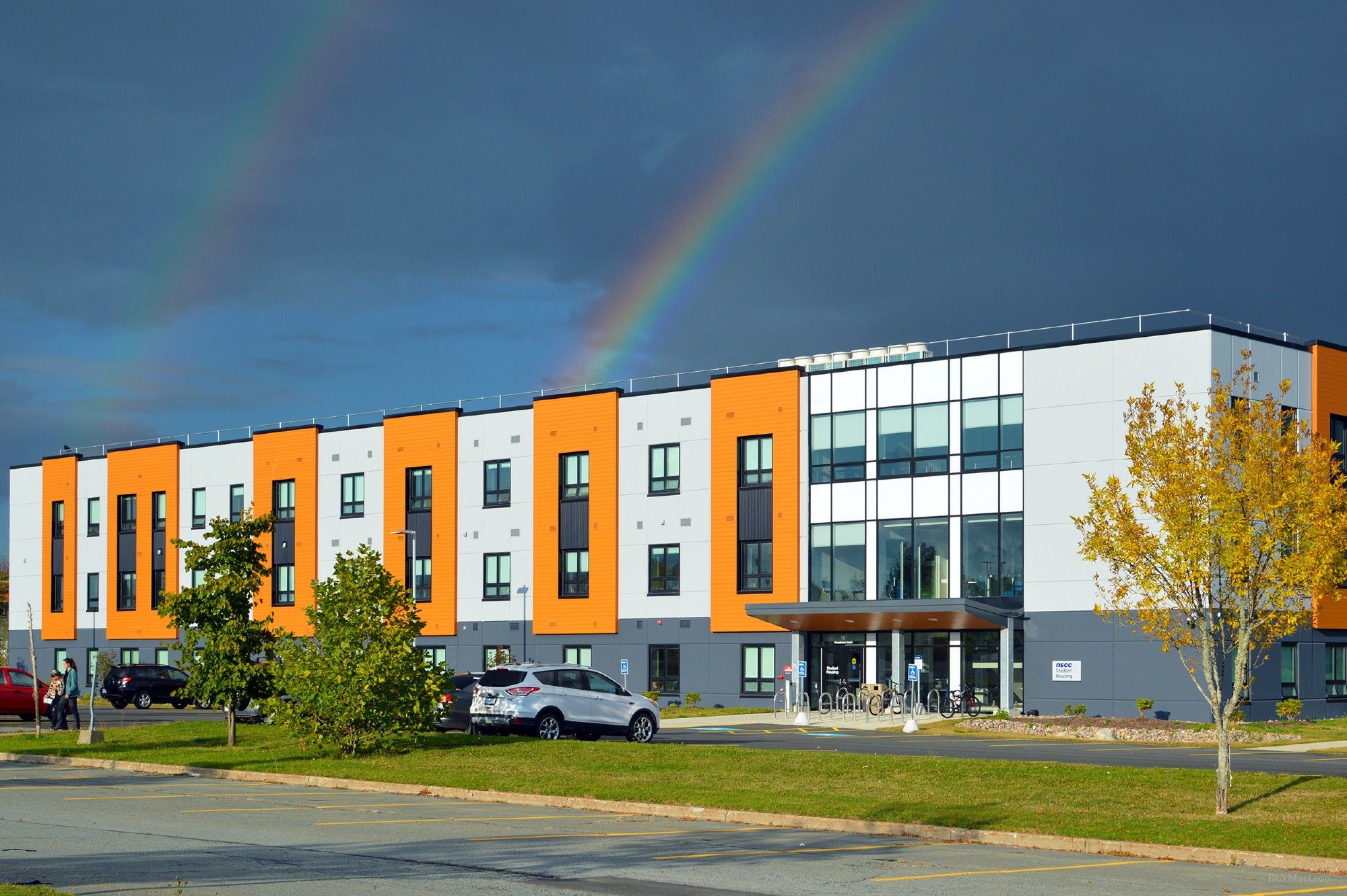 A three-storey, boxy-looking student residence hall next to a mostly empty parking lot. A sign next to the main entrance says "NSCC Student Housing". There is a double rainbow in the sky behind the building.