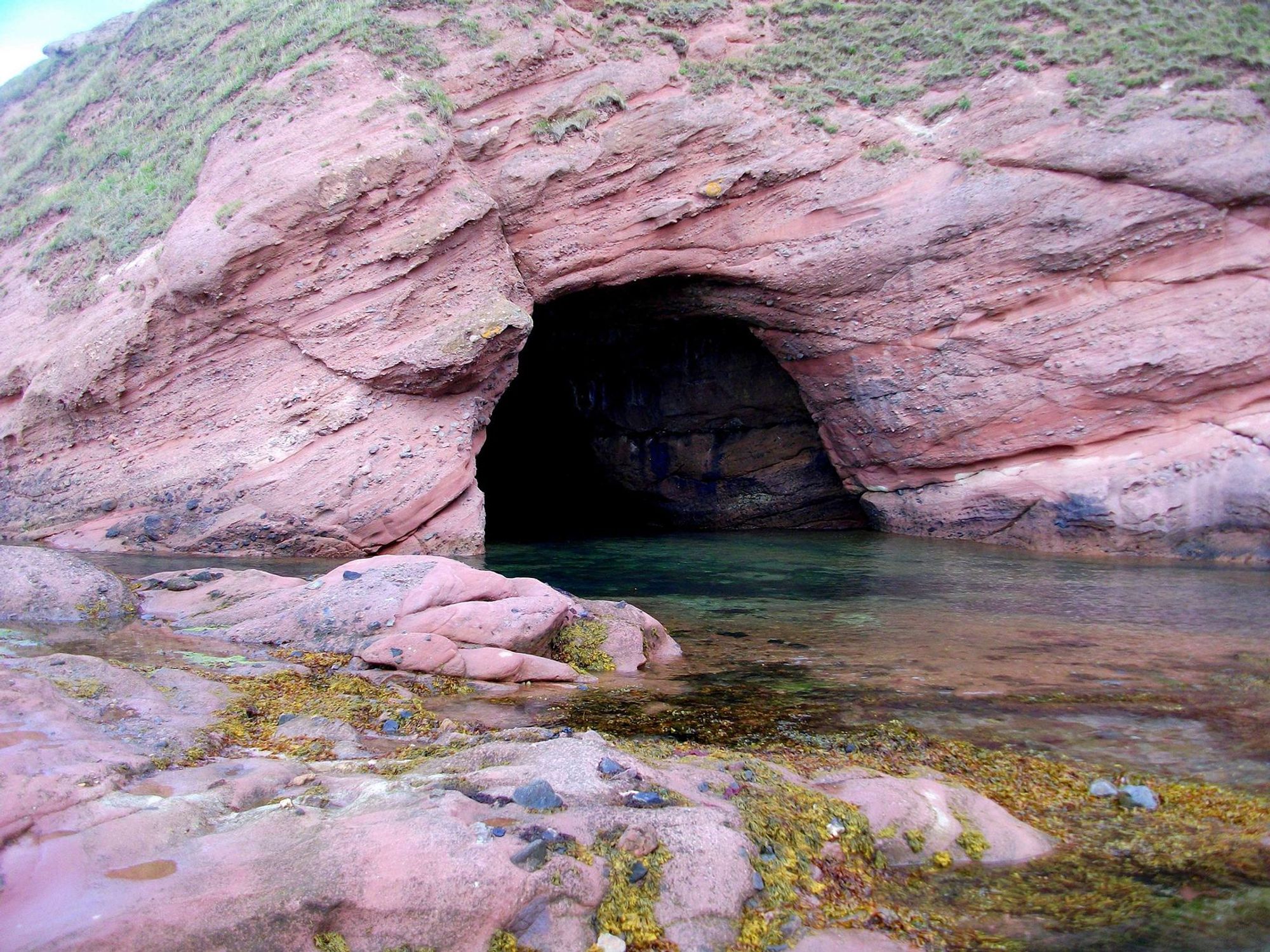 A cave in layered pink rock, fronted by sea-water.