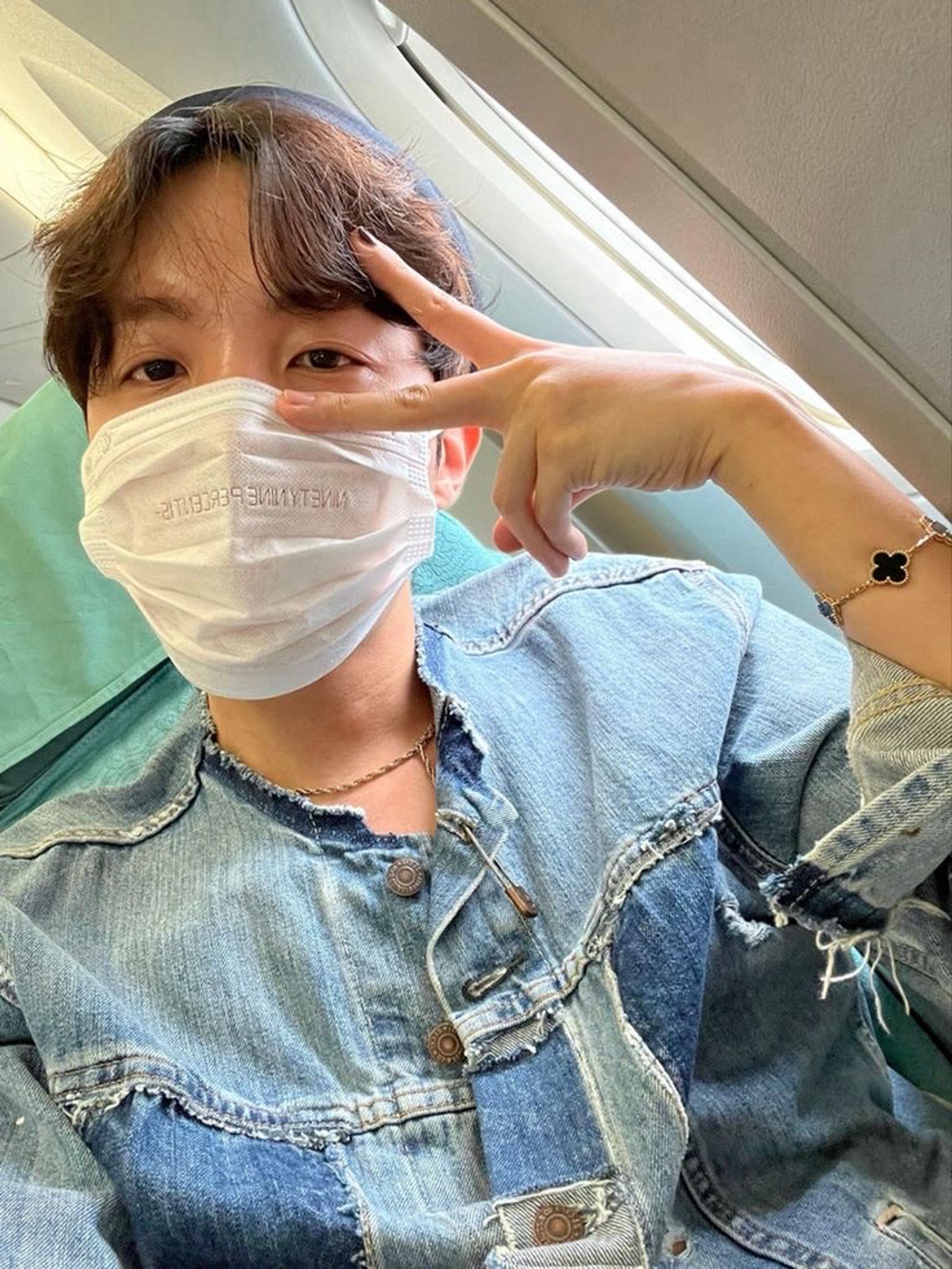 Hobi is seated on an airplane by the window, wearing a white face mask. He has wavy brown hair under a beanie and makes a peace sign with his hand by his face. He is dressed in a blue denim jacket and wears a black clover charm bracelet. Photo was posted by him on Weverse.