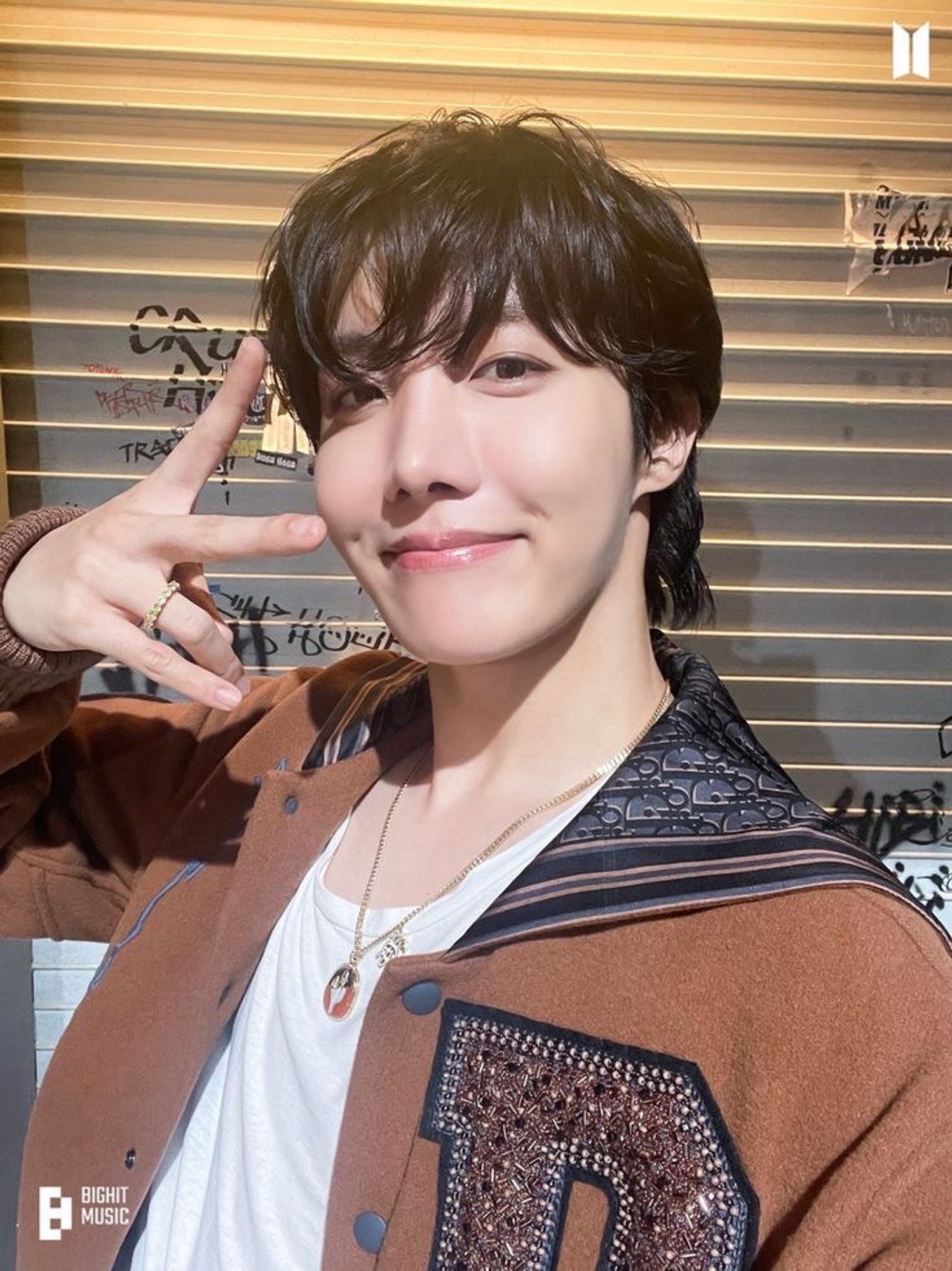 Hobi smiles warmly at the camera and poses with a peace sign. His black hair is slightly tousled, framing his face in soft waves. He wears a stylish brown Dior jacket over a white t-shirt. He also wears a gold pendant necklace and a ring. The background shows a shutter with graffiti covering it. Photo is from ‘Rush Hour (feat j-hope of BTS)’ Photo Sketch.