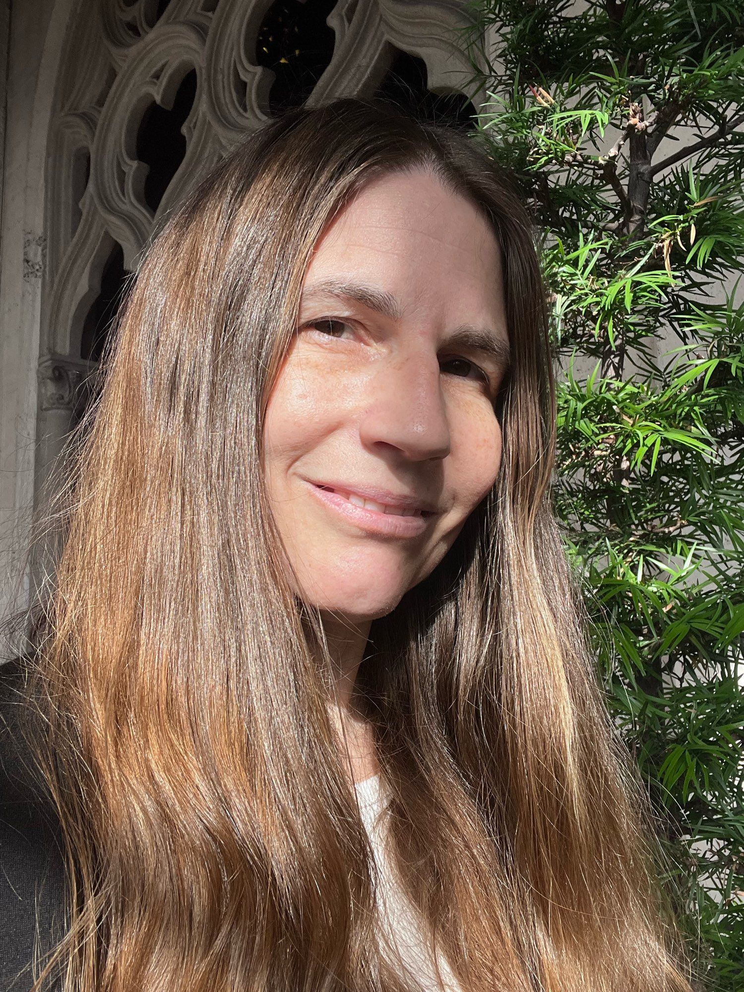 Hobs with hair down, dappled with shadows. Their hair was waist-length and is mostly straight in this photo. Sunlight catches copper tones in their light brown hair.