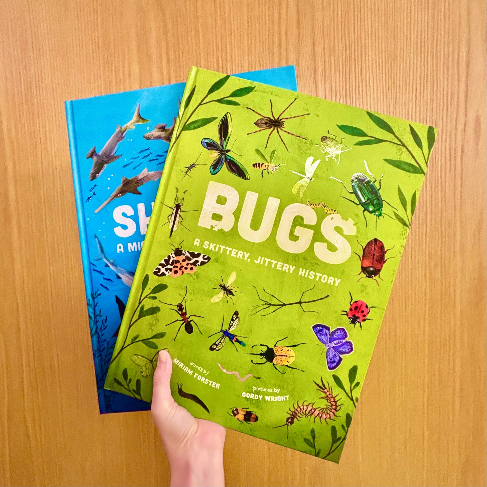 A hand holding two oversized picture books. The back one is blue and called Sharks, a Mighty Bitey History, and the front one is green and titled Bugs, a Skittery Jittery History.