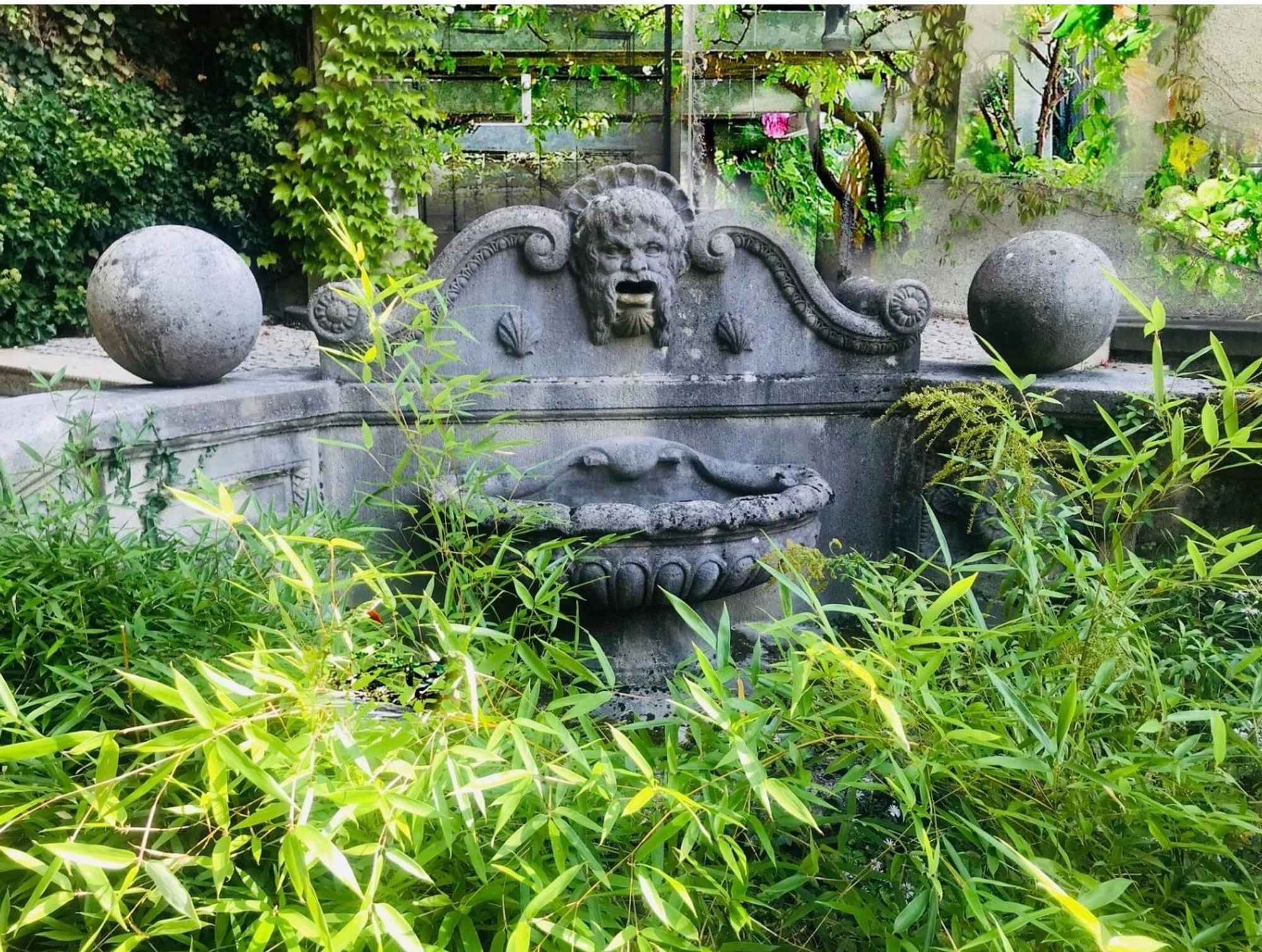 Ein Brunnen aus Stein mit einem verzierten Rand; über dem Wasserbehälter das Relief eines Männer-Gesichts mit Bart, links und rechts davon je eine Muschel.  Auf einer verzierten Balustrade links und rechts eine große Kugel aus Stein.