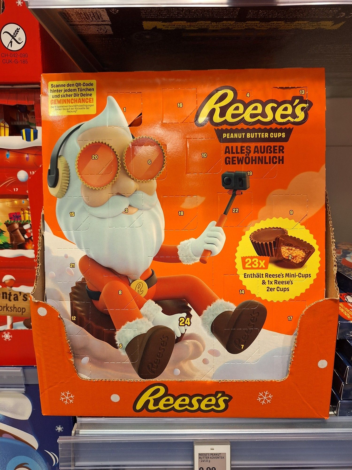 Ein oranger Adventskalender mit nichts als Reese's Peanut Buttercups gefüllt. Darauf ist ein Weihnachtsmann abgebildet, der auf einem Buttercup als Schlitten einen Berg aus Milch(?) herunterrutscht. Er hat eune Kamera mit Selfiestick in der Hand, Kopfhörer in Buttercup-Form und gruselige Buttercup-Augen.