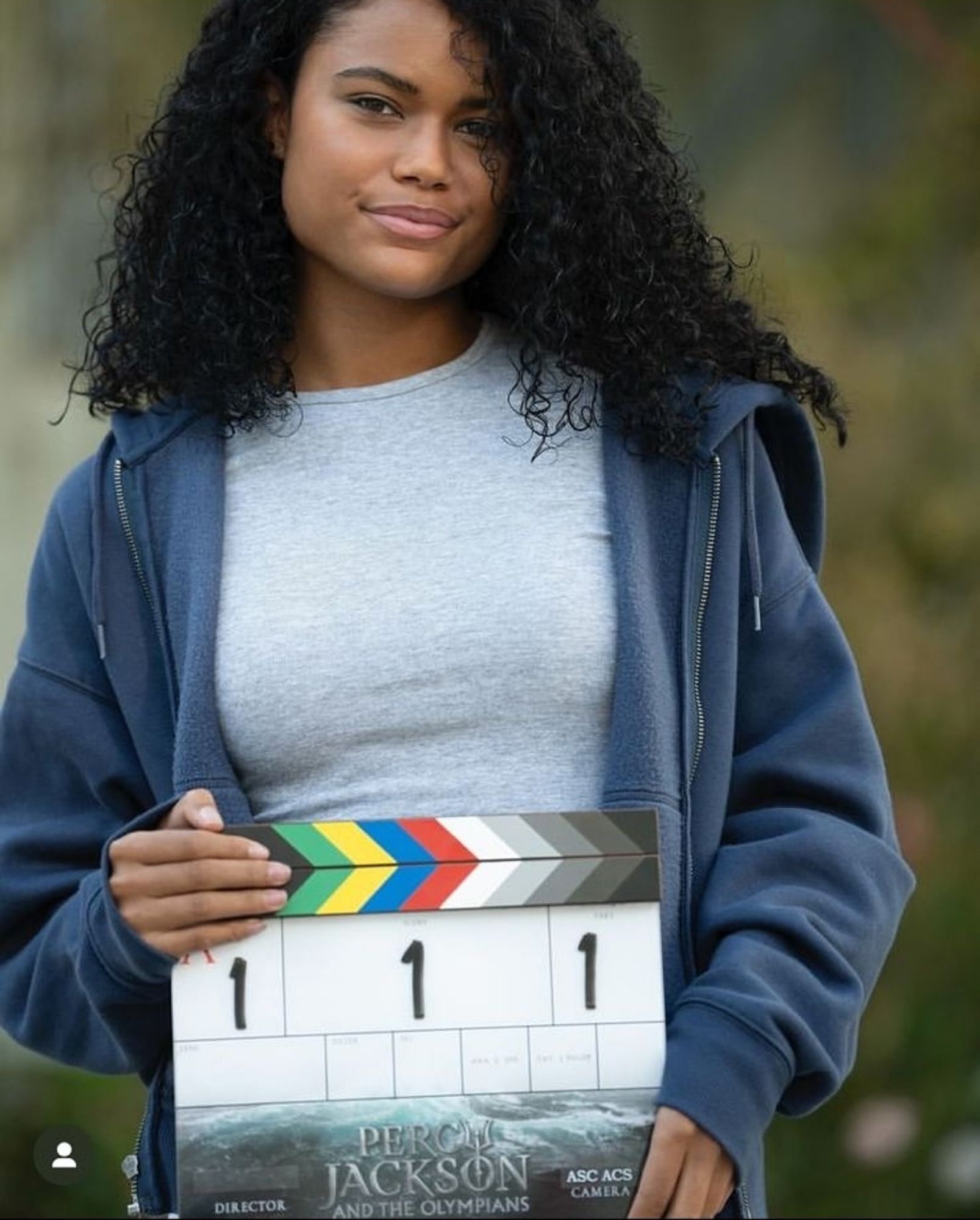 Foto de Tamara Smart no set de Percy Jackson e os Olimpianos. A atriz, uma jovem negra de cabelo cacheado preto, segura uma claquete da série e usa uma blusa cinza e um casaco de moletom azul marinho.