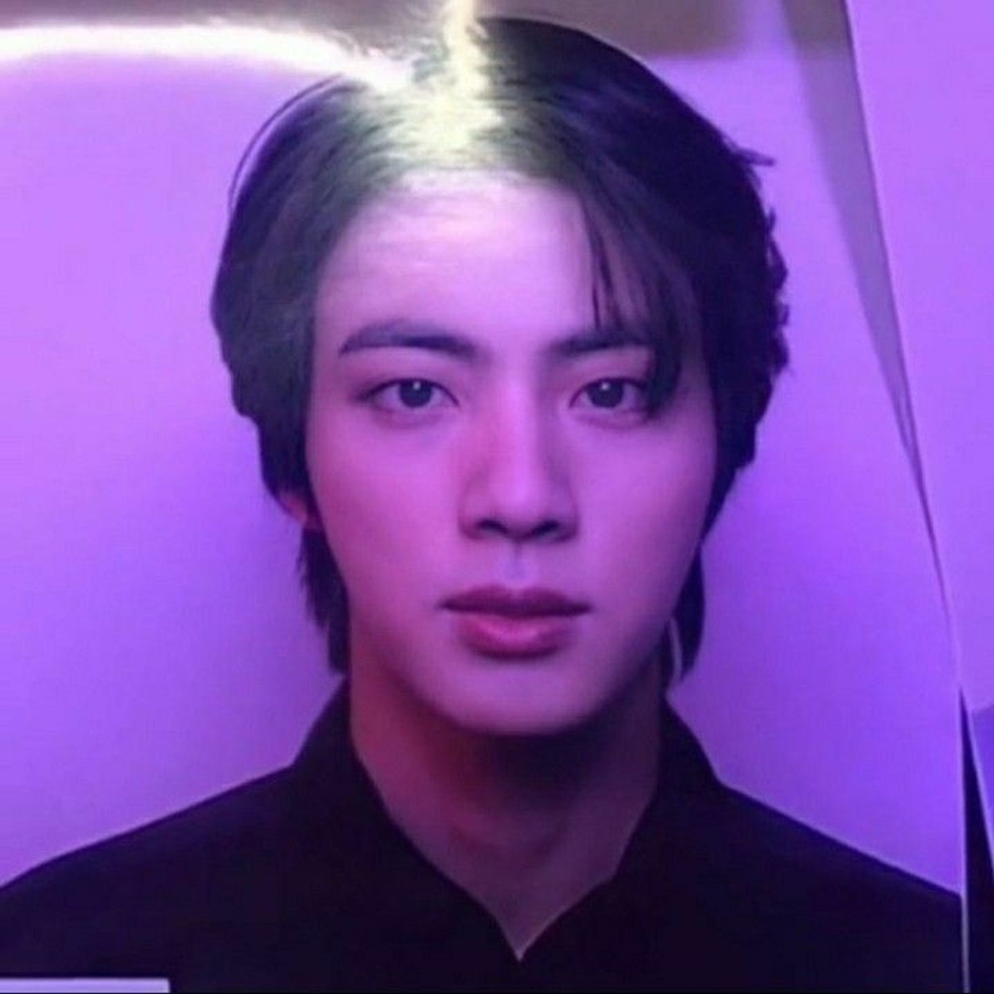 The image shows a close-up photo of Jin from BTS, which appears to be from James Corden’s photowall. His face is illuminated with a purple-tinted light, highlighting his soft features and clear complexion. He has a calm and composed expression, with his dark hair framing his face. The lighting gives the photo a slightly ethereal and dramatic effect.