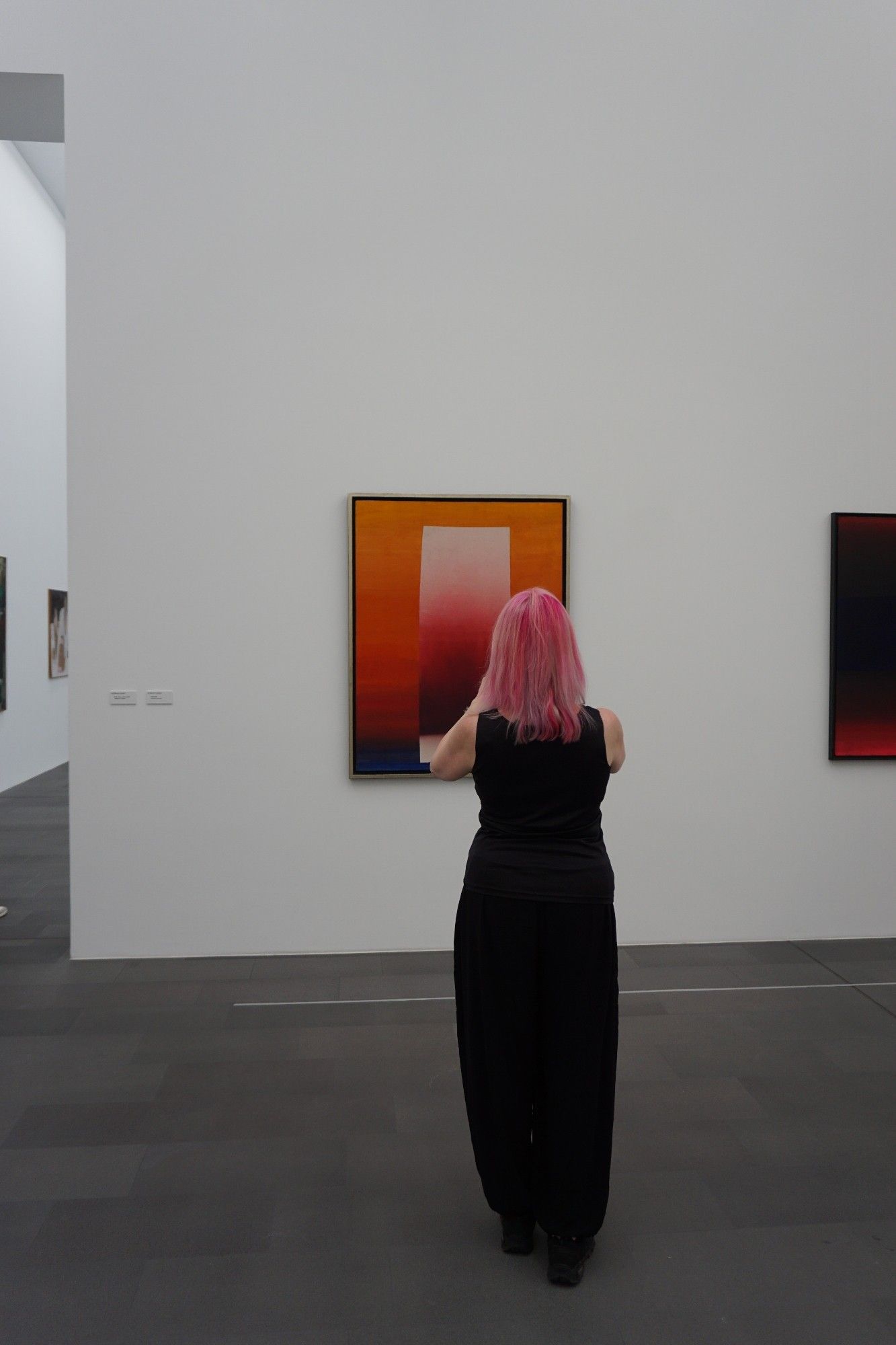 Frau in schwarzer Kleidung mit pinken Haaren steht in einer Galerie vor einem Bild mit orange-rot-pinken Farbverläufen