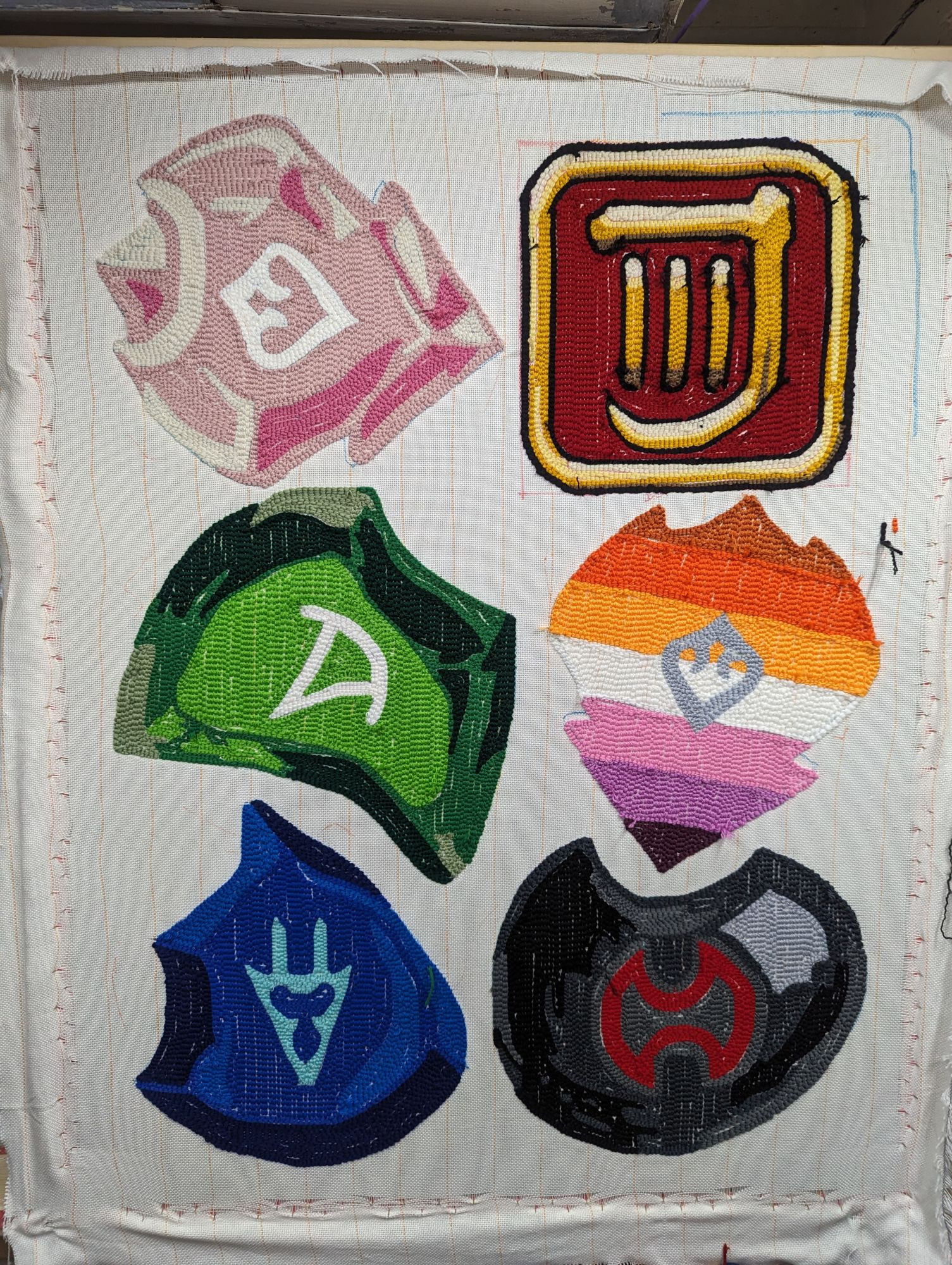 A picture of six rugs on a rug tufting frame, still part of the main tufting cloth before they're glued and cut apart. The rugs are a Dancer Soul Crystal, backwards Bard Job Symbol, a Summoner Soul Crystal, a lesbian pride flag Dancer Soul Crystal, a Dragoon Soul Crystal, and a black Warrior Soul Crystal.