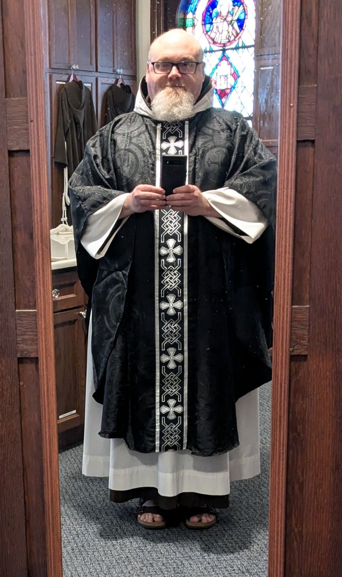 sacristy mirror selfie of a middle-aged Capuchin priest vested for Mass with a gothic-style black chasuble