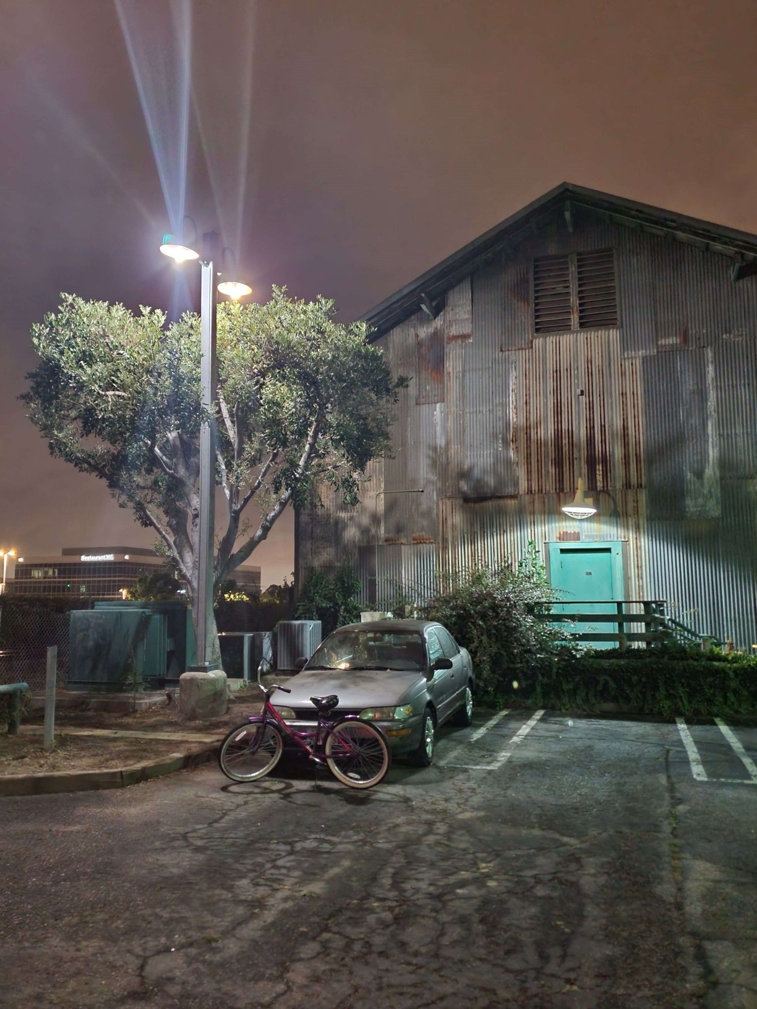 An otherworldly picture of a run-down warehouse, car, and bike, and tree at midnight exuding strong Americana vibes. The teal door matches the teal industrial machines and bikes in frame. A light post illuminates the scene.