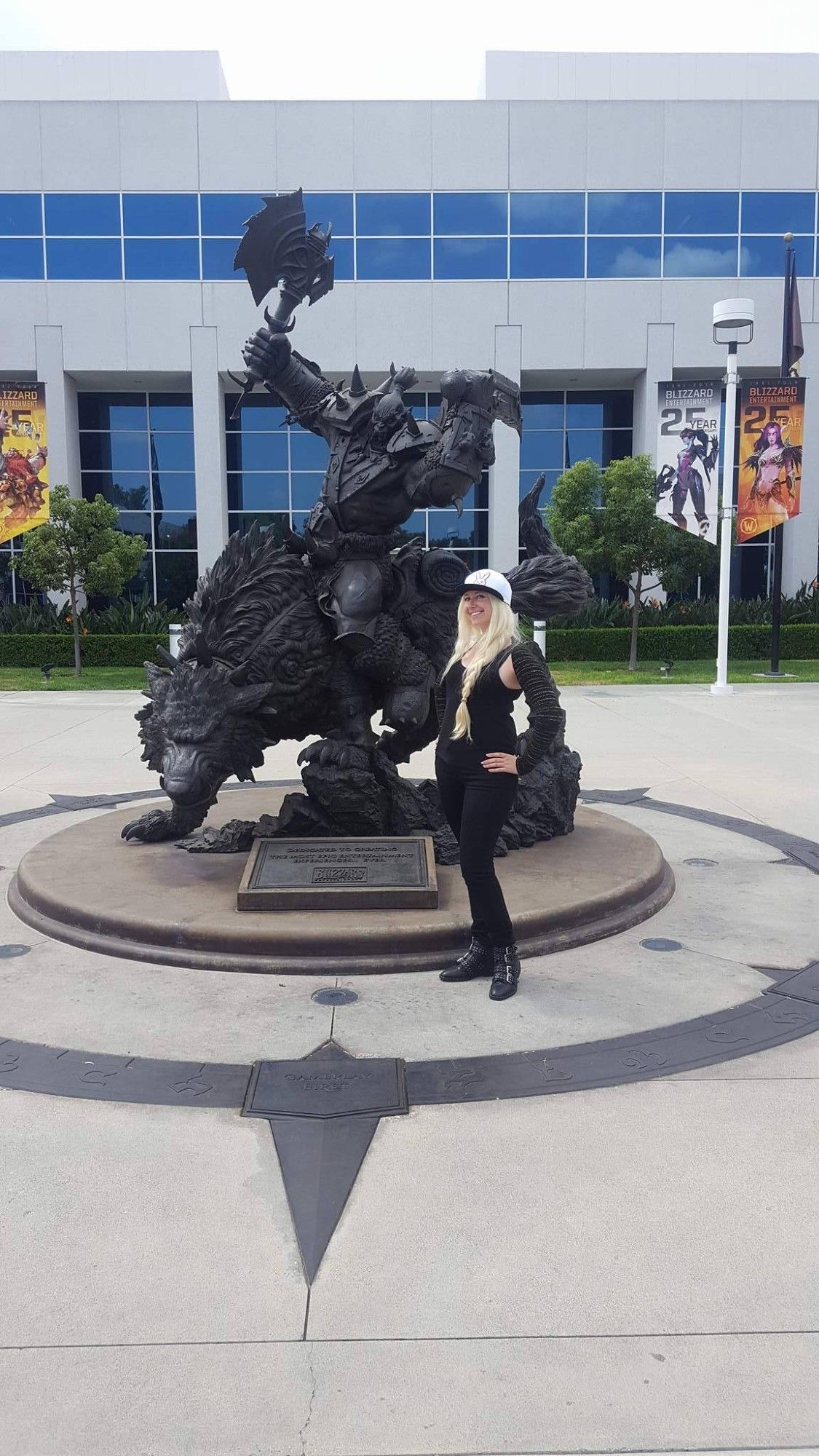 The author Katie stands in front of the Blizzard Campus Orc Statue. She has long, blonde hair braided to the side adorned by a D.Va bunny logo hat. Her outfit includes a black ribbed tank top with a black and gold shrug that shows off her shoulders, black pants, and black leather shoes covered in shiny silver buckles and studs.