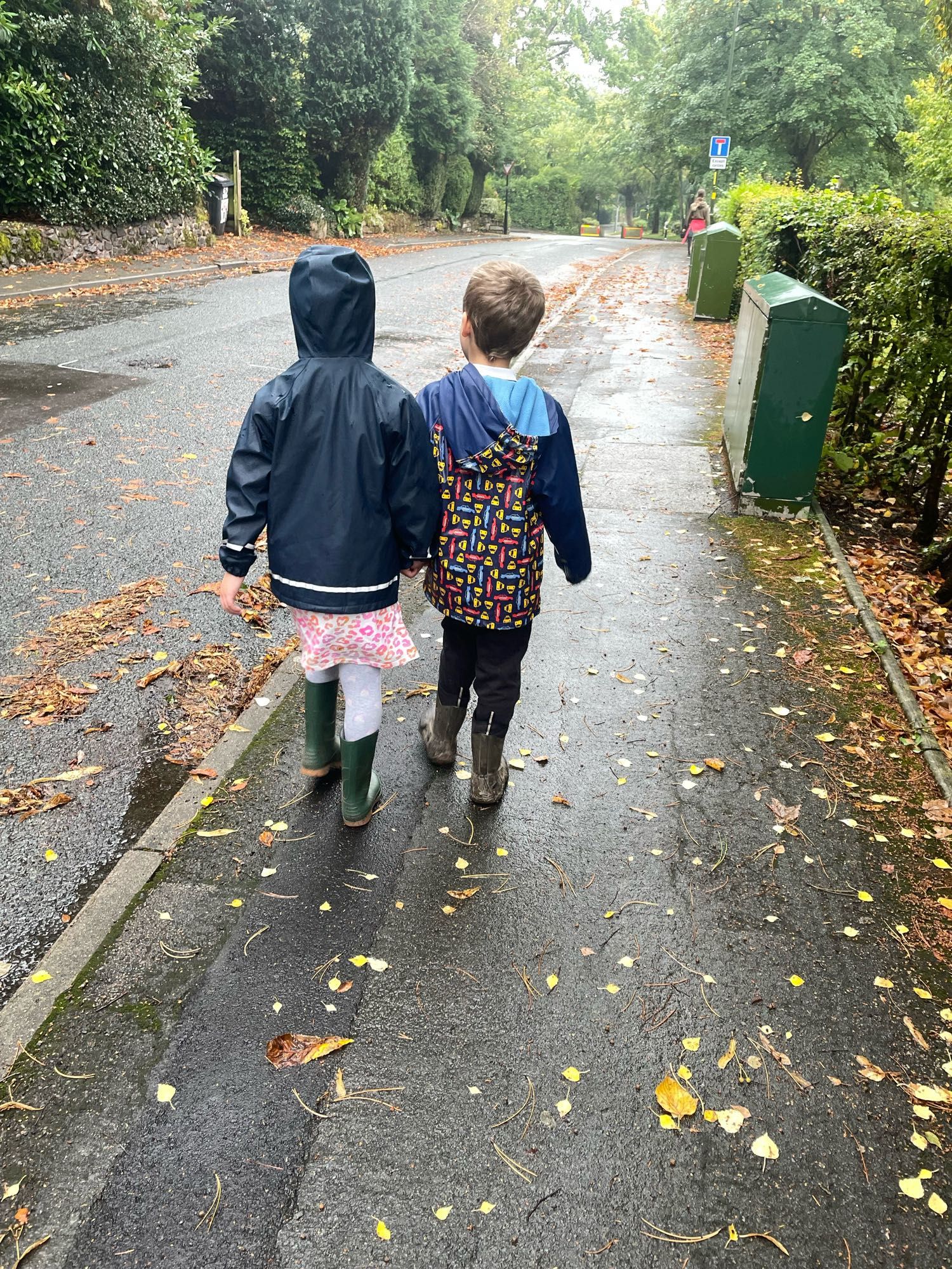 Two children walking on a pavement with wellies and rain coats on. It’s raining and the ground is wet.