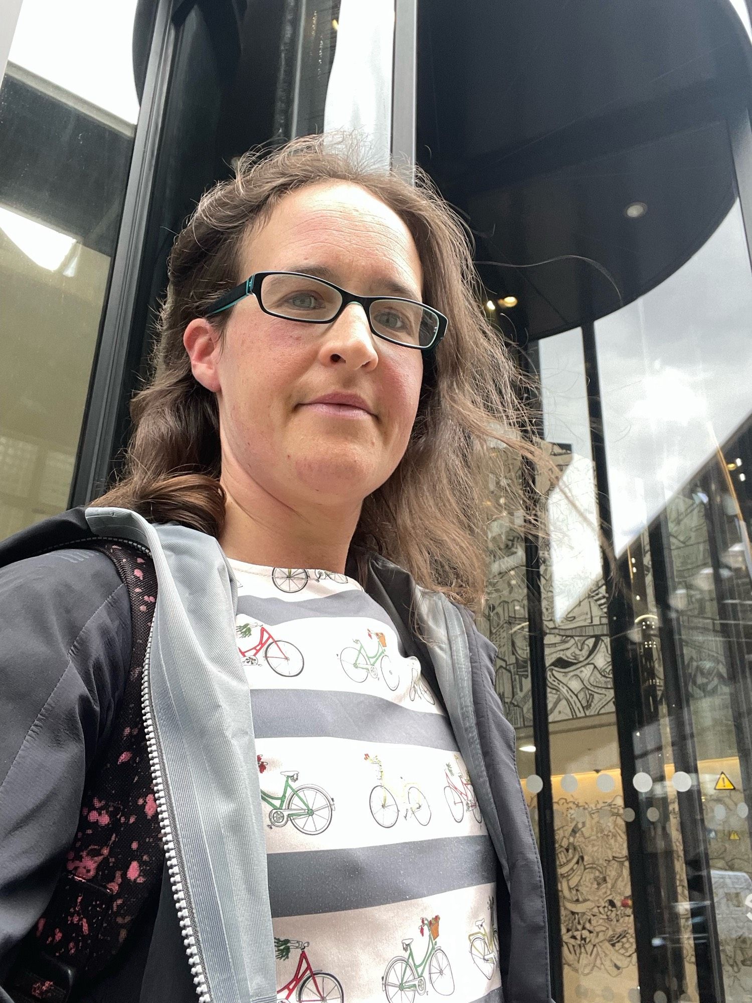 A selfie of me at the revolving doors of Google HQ London. I’m wearing a dress with bikes on and a black rain coat.