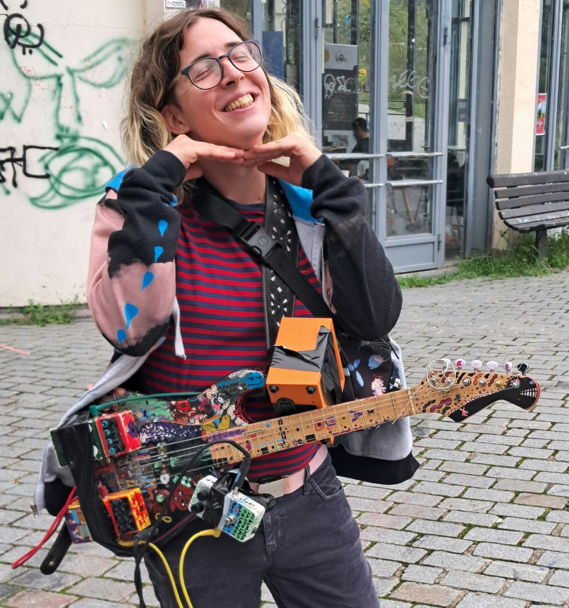 Rose is smiling, outside, holding a guitar were effect pedals and amp are tapped on the guitar.