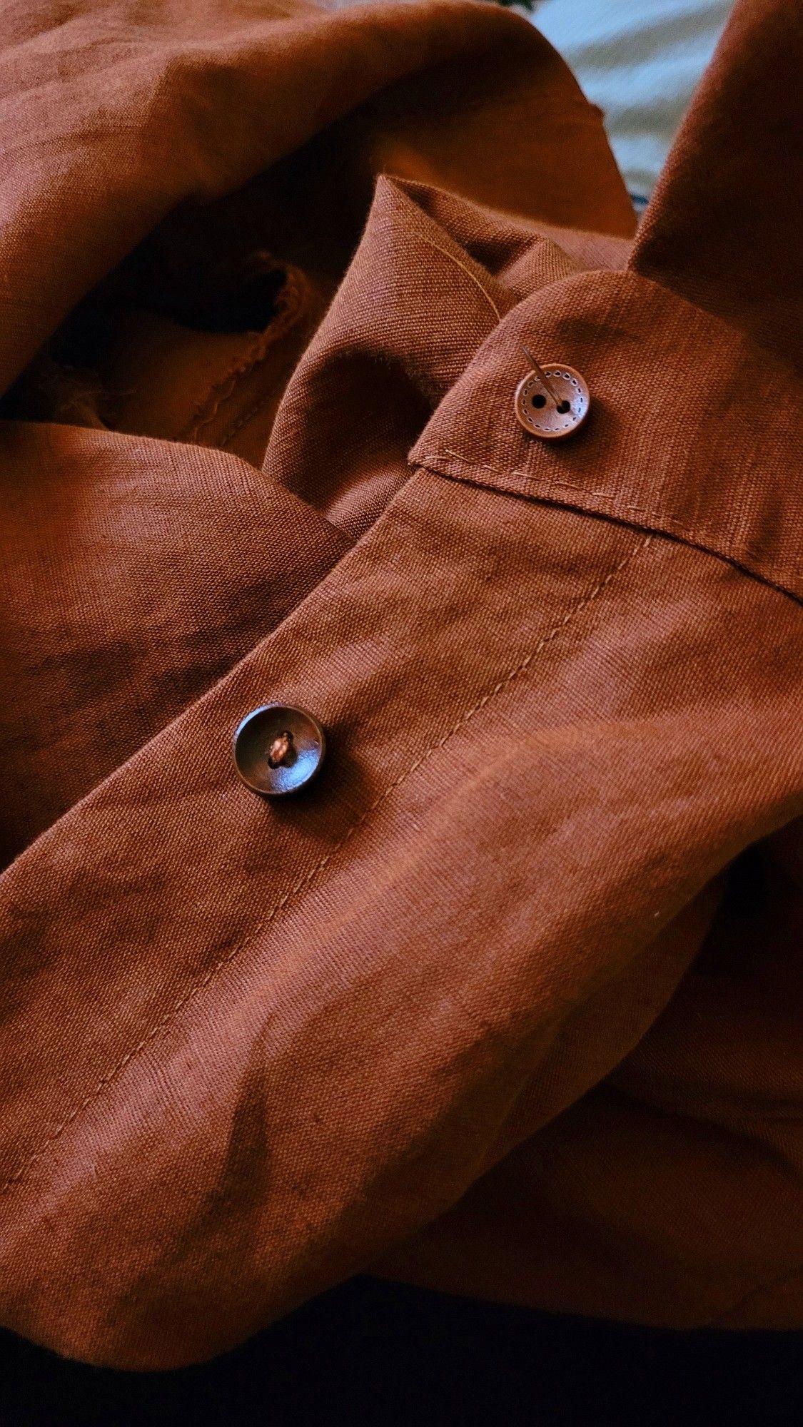 close up of a work in progress linen shirt. it shows buttons being handstitched. the colour is a warm rust.