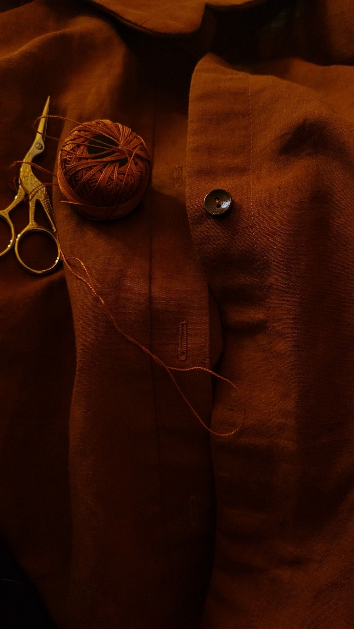 close up of a work in progress linen shirt. it shows the progress of buttons being hand stitched. the colour is a very dark warm rust in dim lighting.