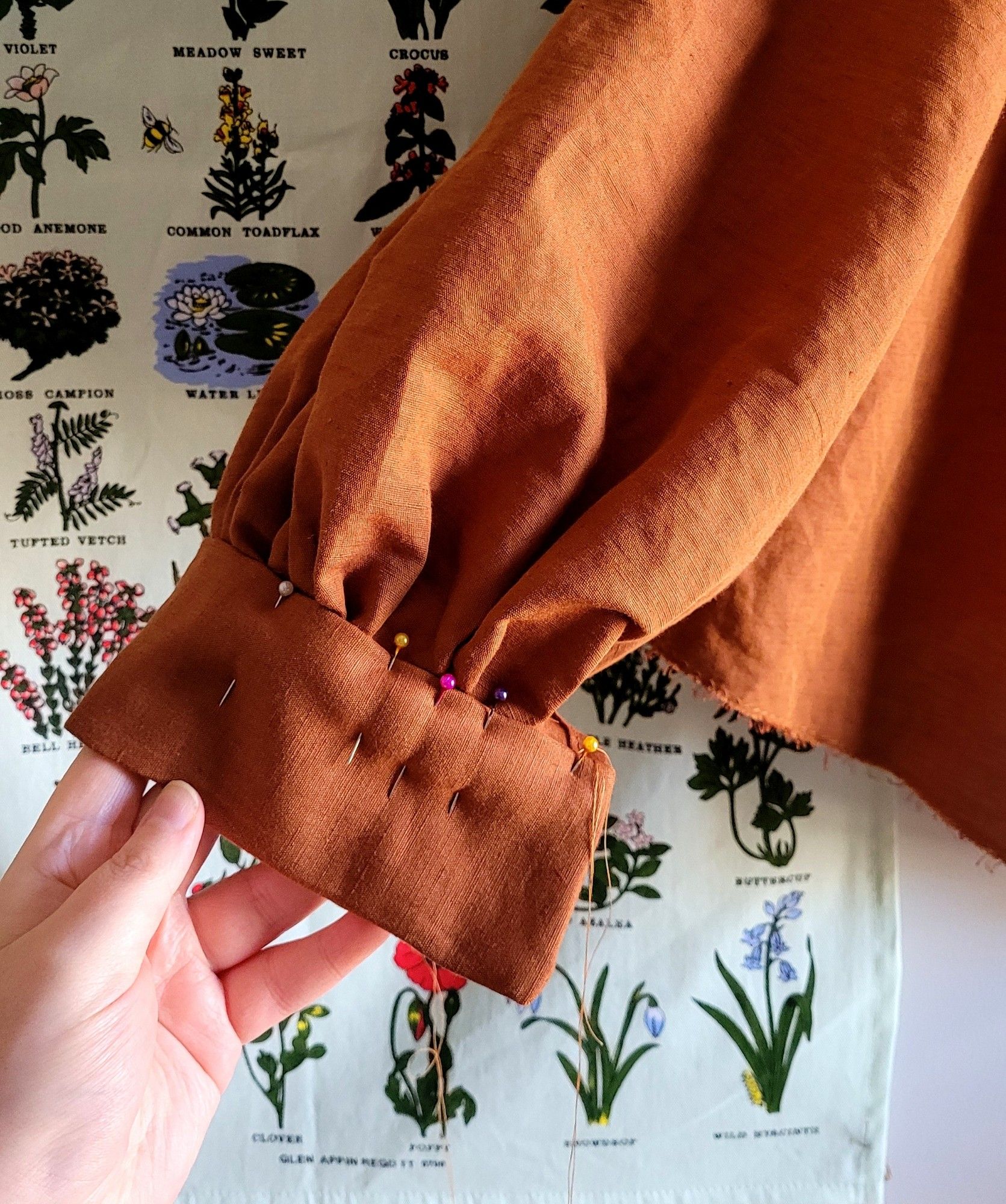 close up of a work in progress linen shirt. it shows the cuffs being attached with sewing pins to a gathered sleeve. the colour is a warm rust.